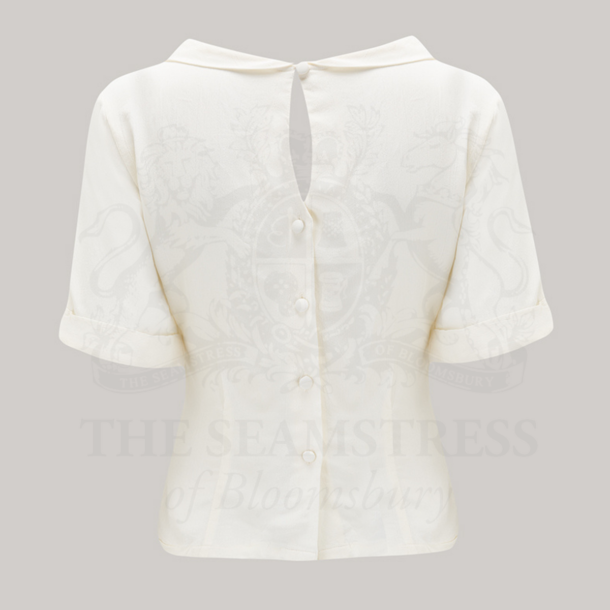 A 1940s style short sleeve cream blouse. Featuring a side tie-neck collar and button fastenings down the back of the blouse.
