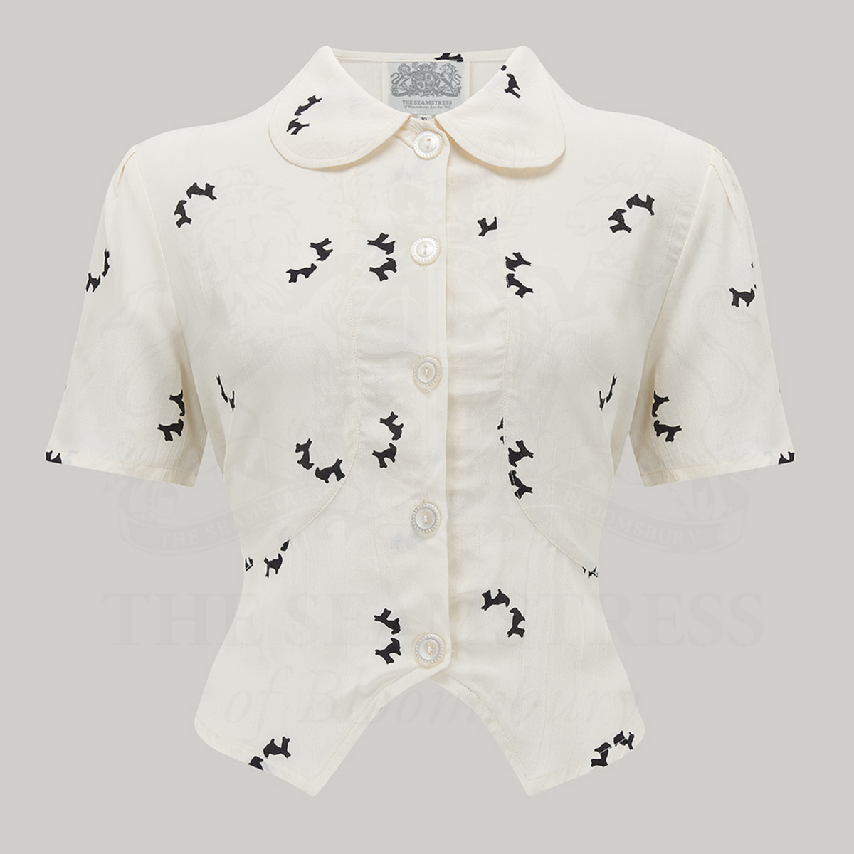 1940s style short sleeve blouse. The blouse is cream coloured with small black dogs dotted around the blouse. Featuring a Peter-Pan front collar, buttons down the front, and a tie-waist belt. 