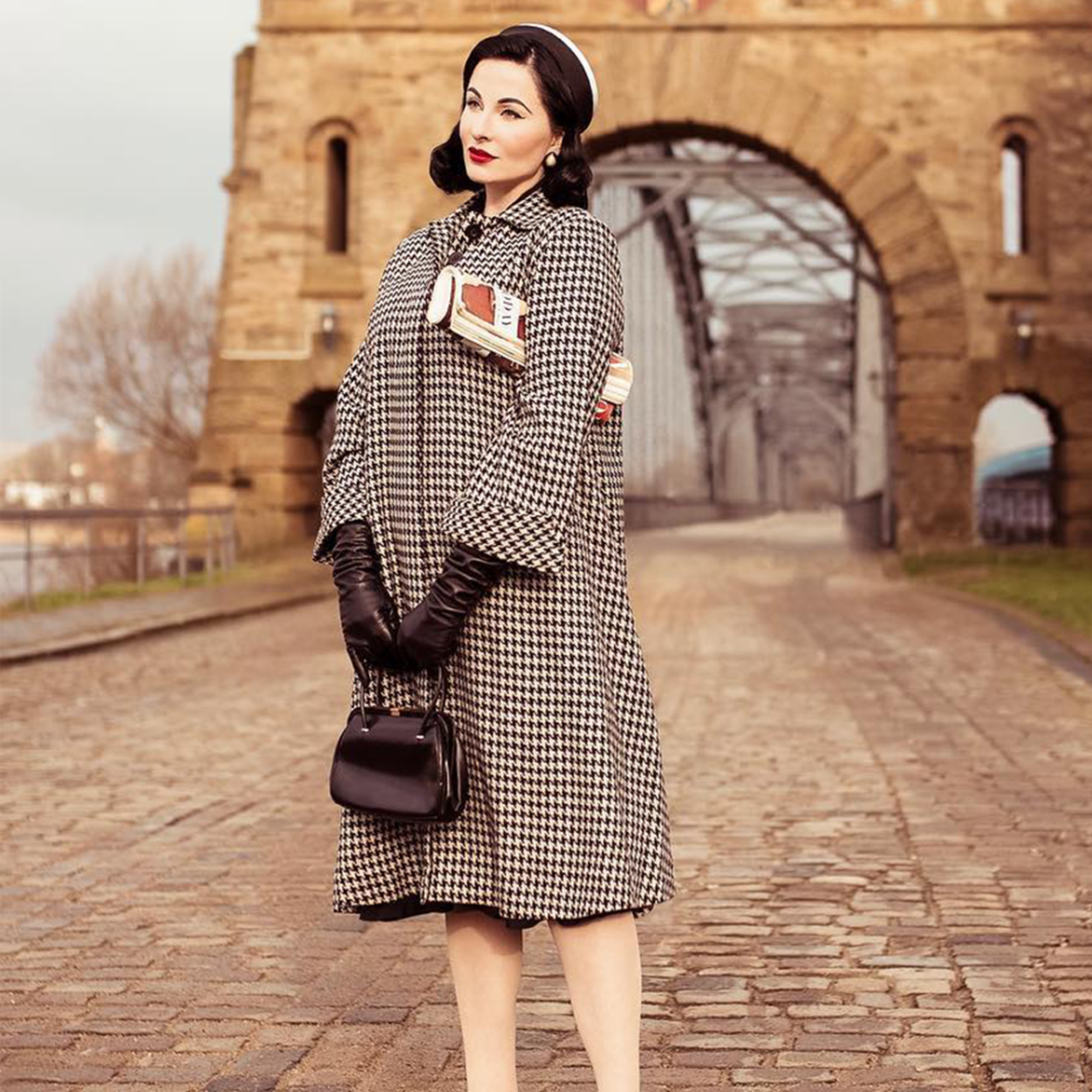 Model wears 1940s style 43 inch long swing jacket. The jacket is in black and white houndstooth print, has three buttons at the top front, small shoulder pads, and is lined in satin. Model has paired with a black bag and gloves, and has styled 1940s makeup and hair. 