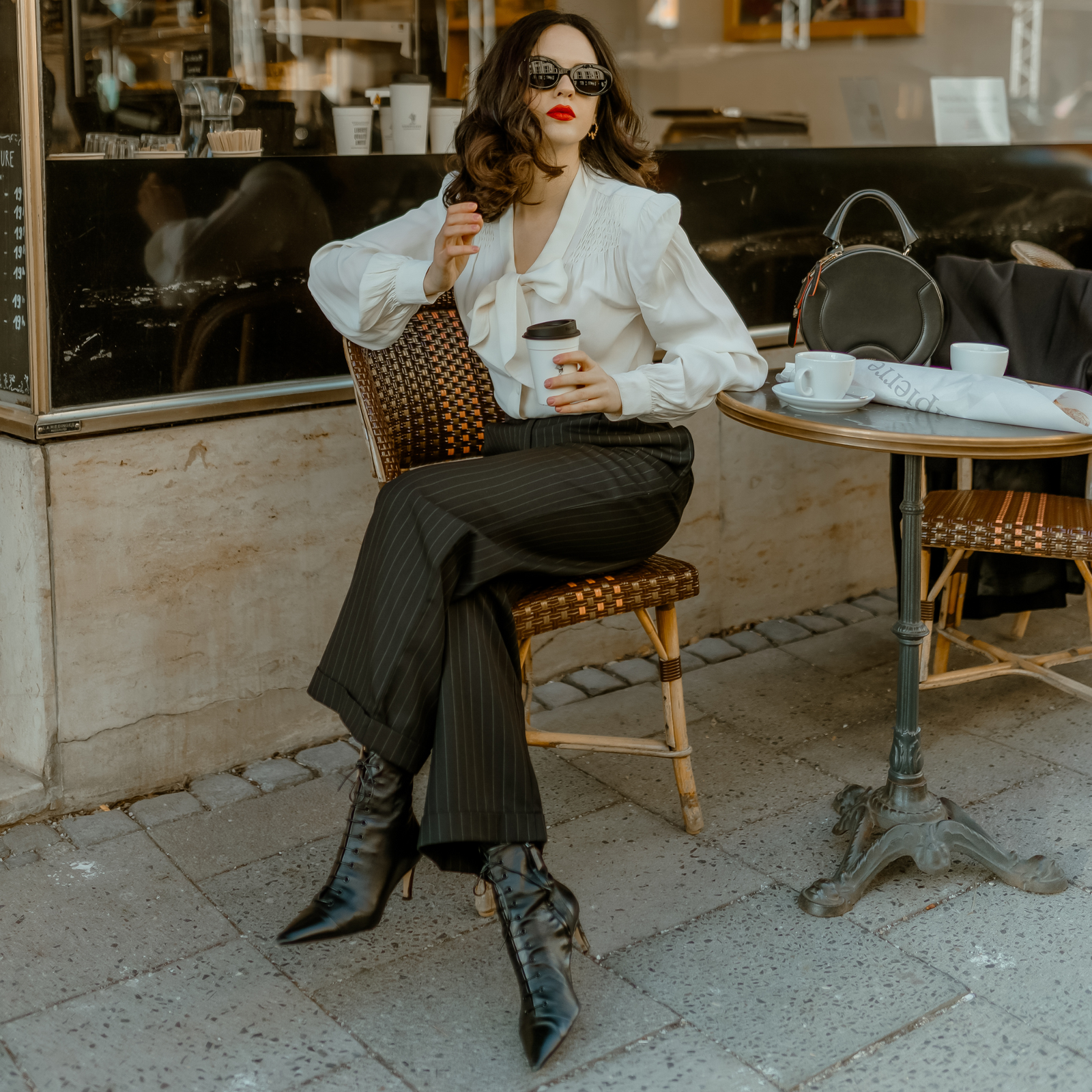 A person seated at an outdoor cafe wearing high-waisted black pinstripe tailored trousers with side pockets and turn-ups, paired with a white blouse and black boots.