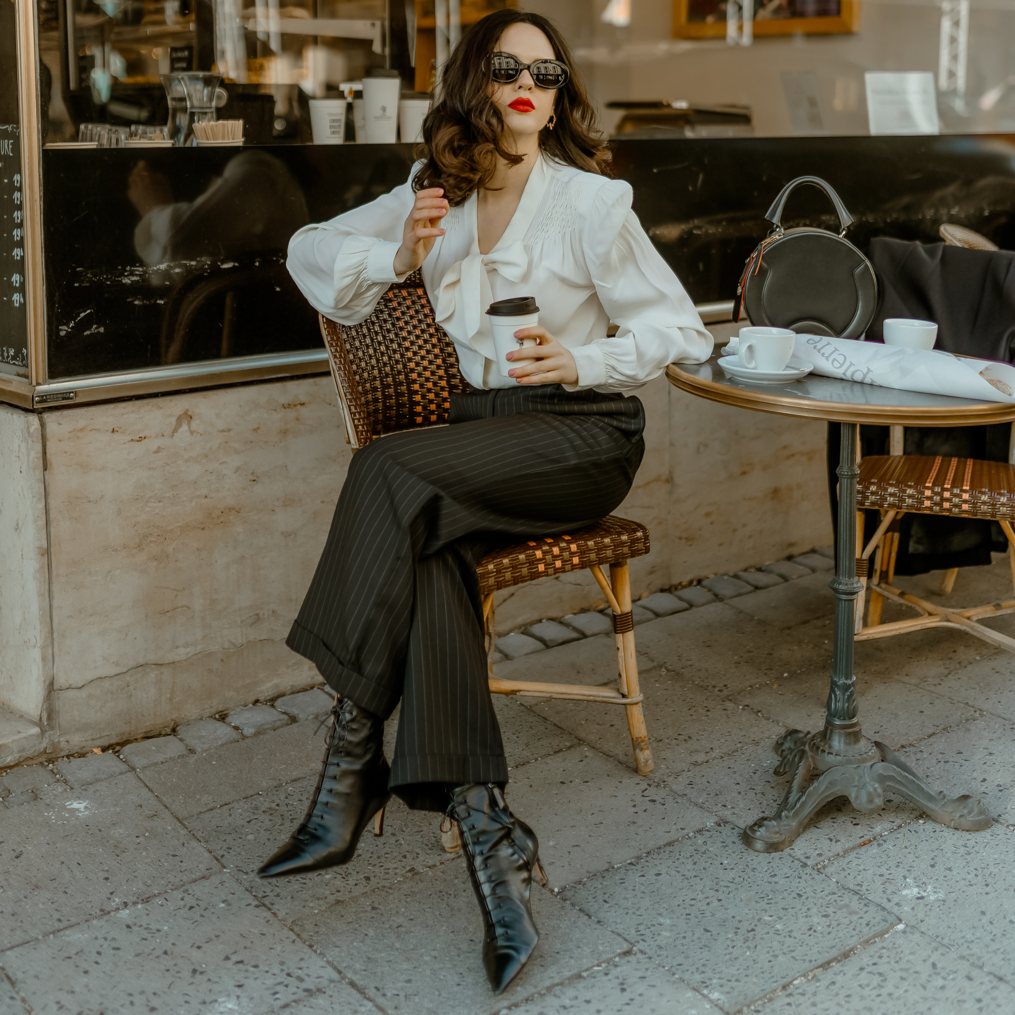 A person seated at an outdoor cafe wearing high-waisted black pinstripe tailored trousers with side pockets and turn-ups, paired with a white blouse and black boots.