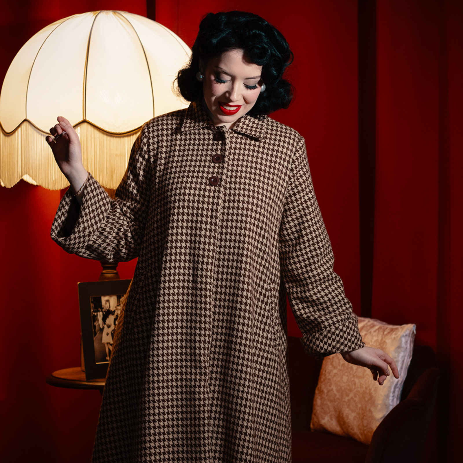 A woman in a houndstooth coat is standing in a room with a lamp and a red wall.
