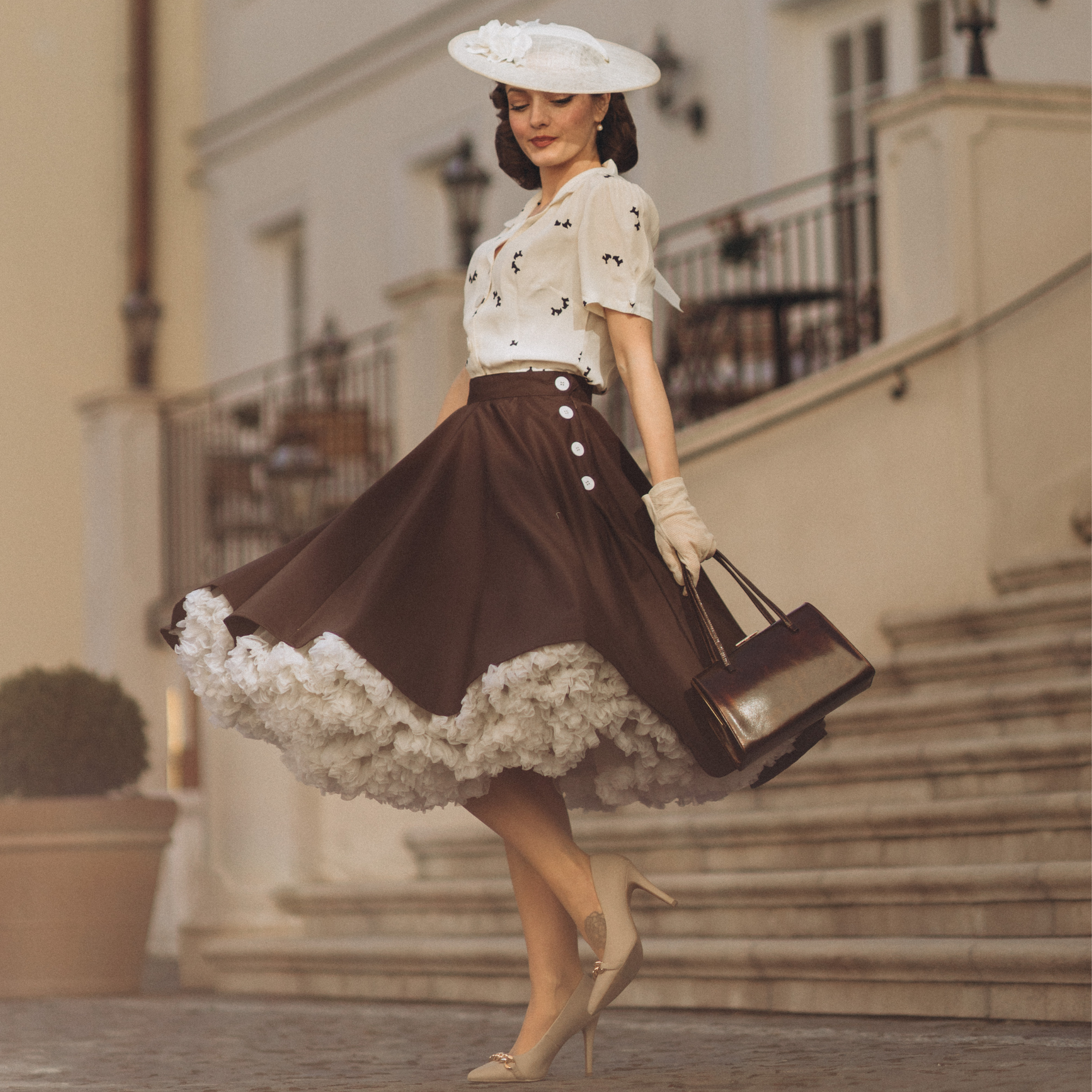 A woman wearing a brown swing dance skirt with decorative buttons, paired with a white top and a white hat, standing in front of a building with stairs.