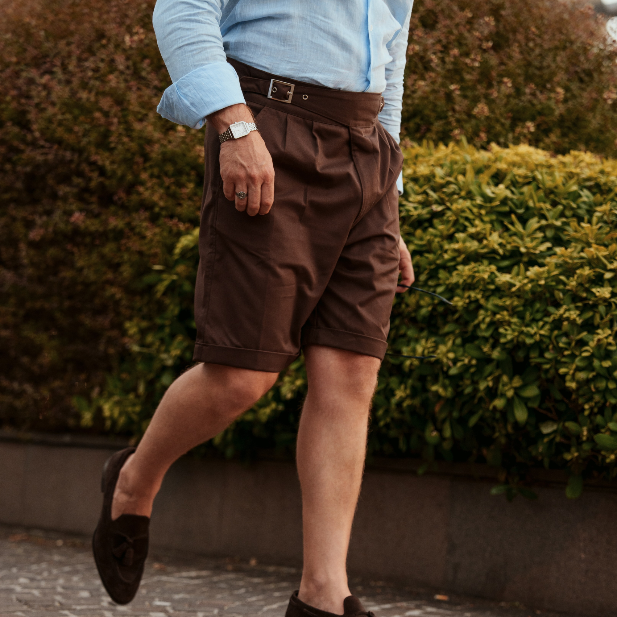 A man wearing brown pleated shorts, a light blue button-down shirt, and dark loafers walking outdoors in front of green hedges.