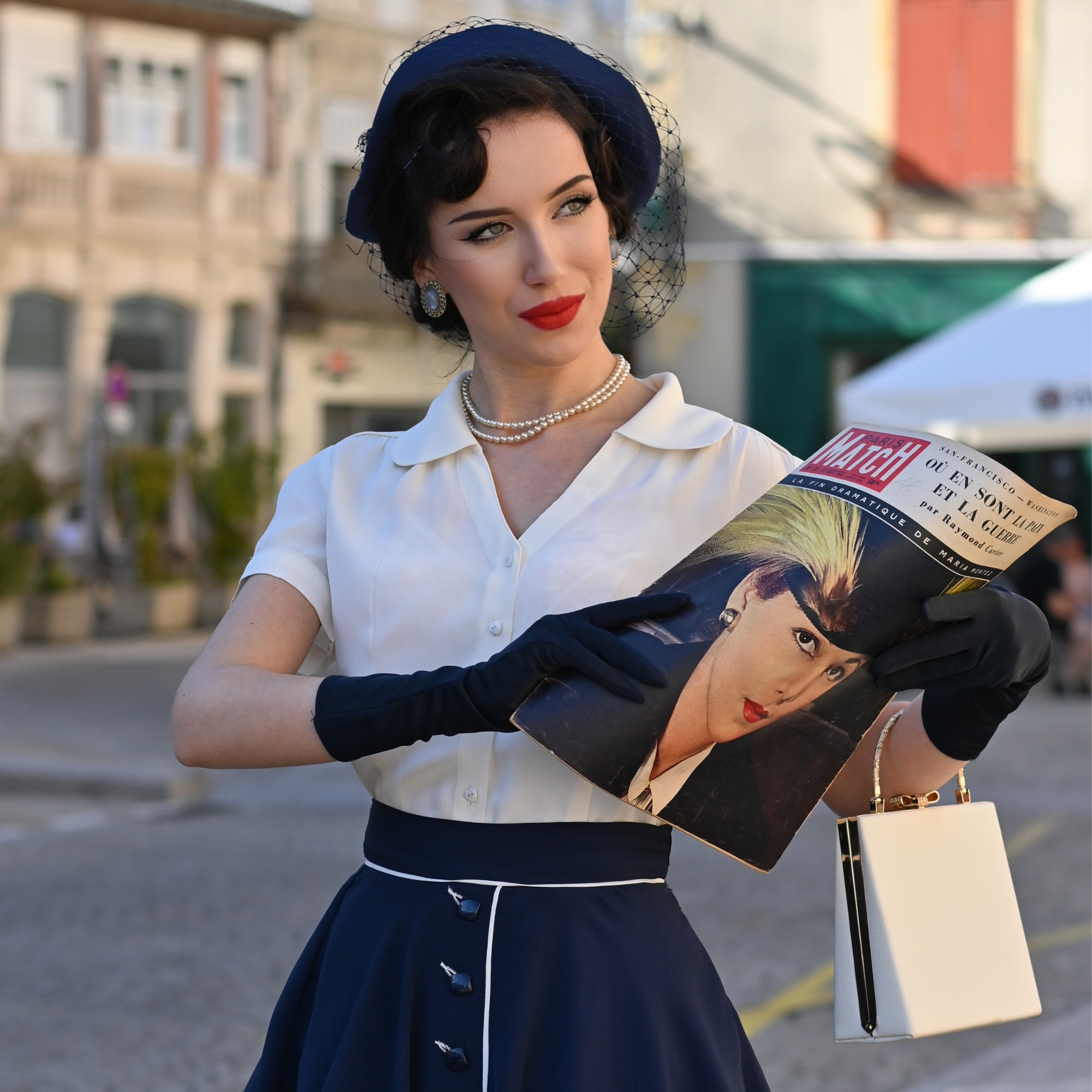 A person wearing a vintage-style cream blouse with a Peter Pan-style collar, puff sleeves, and a fitted waist, paired with a navy skirt, holding a magazine and wearing gloves.
