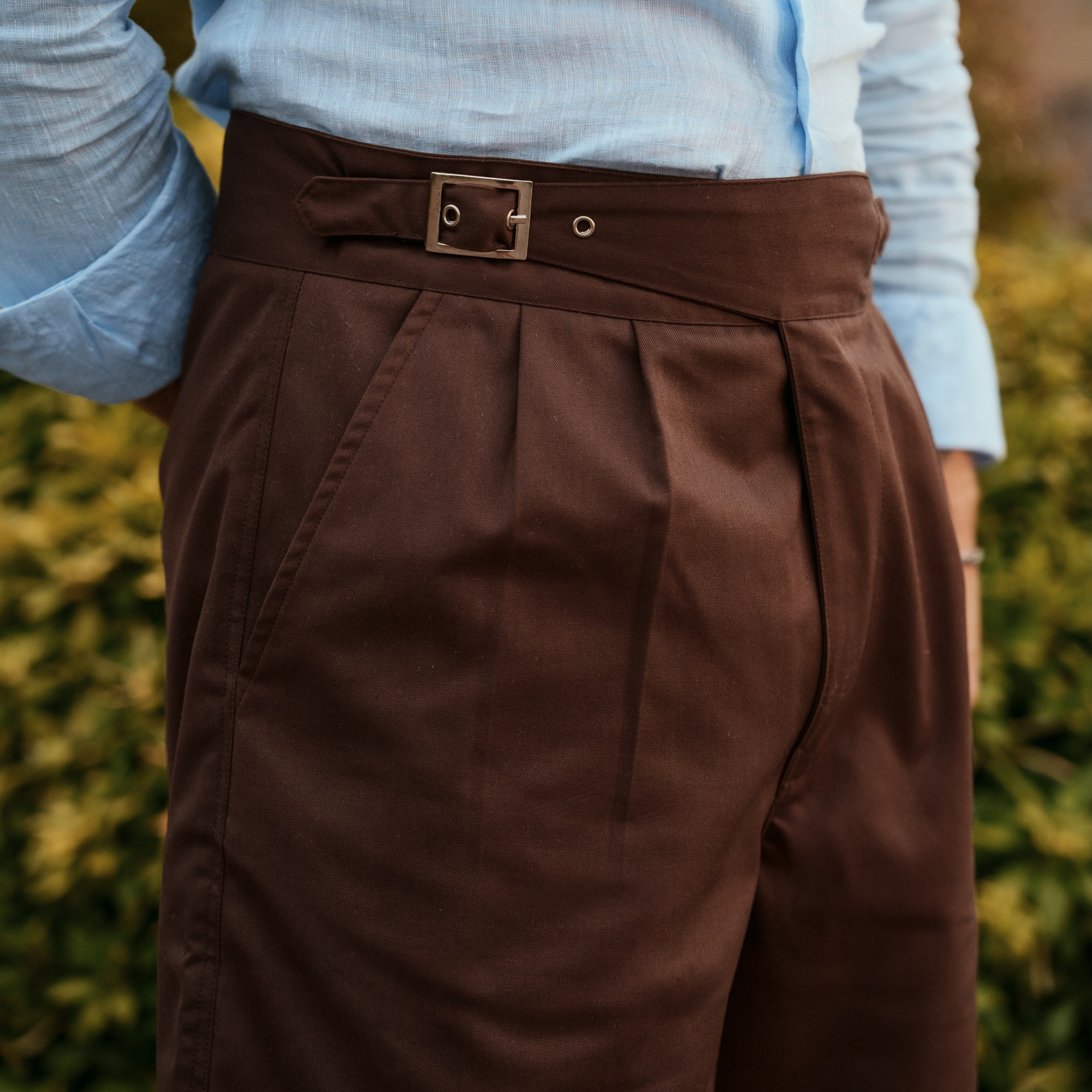Close-up of a person wearing dark brown pleated shorts and alight blue shirt, standing outdoors with blurred green foliage in the background.