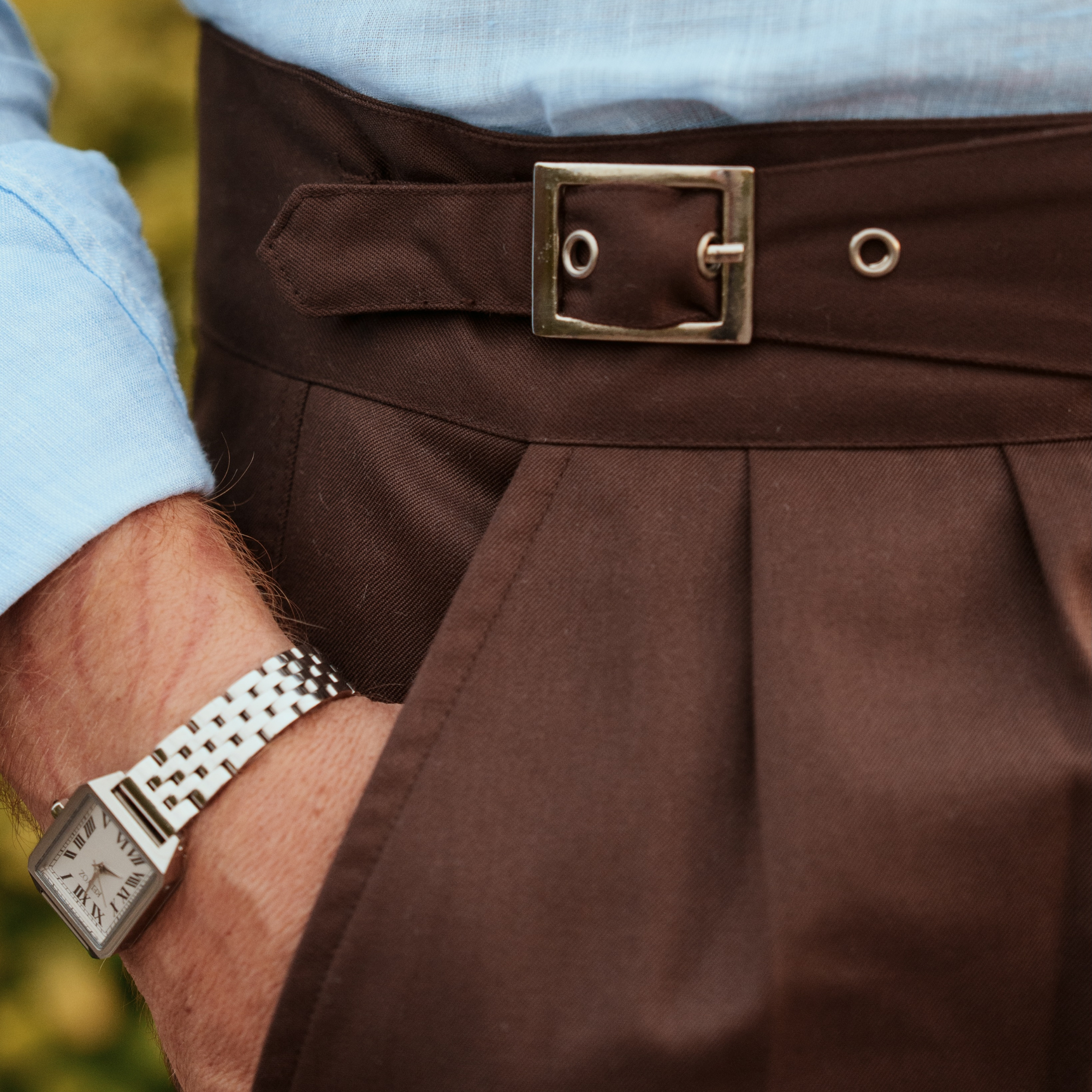 Close-up of a person wearing dark brown pleated shorts featuring a square metal buckle, paired with a light blue dress shirt and a silver watch.