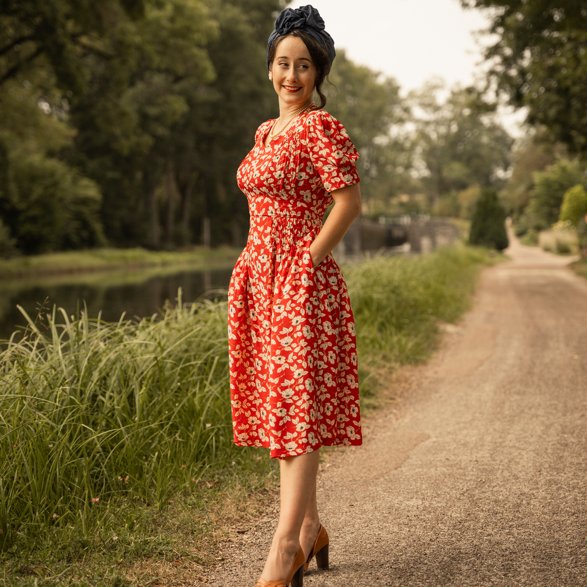 A 1940s-style short-sleeve dress in red with floral design. Featuring a sweetheart neckline, smocking detailing, pockets, and a waist tie.
