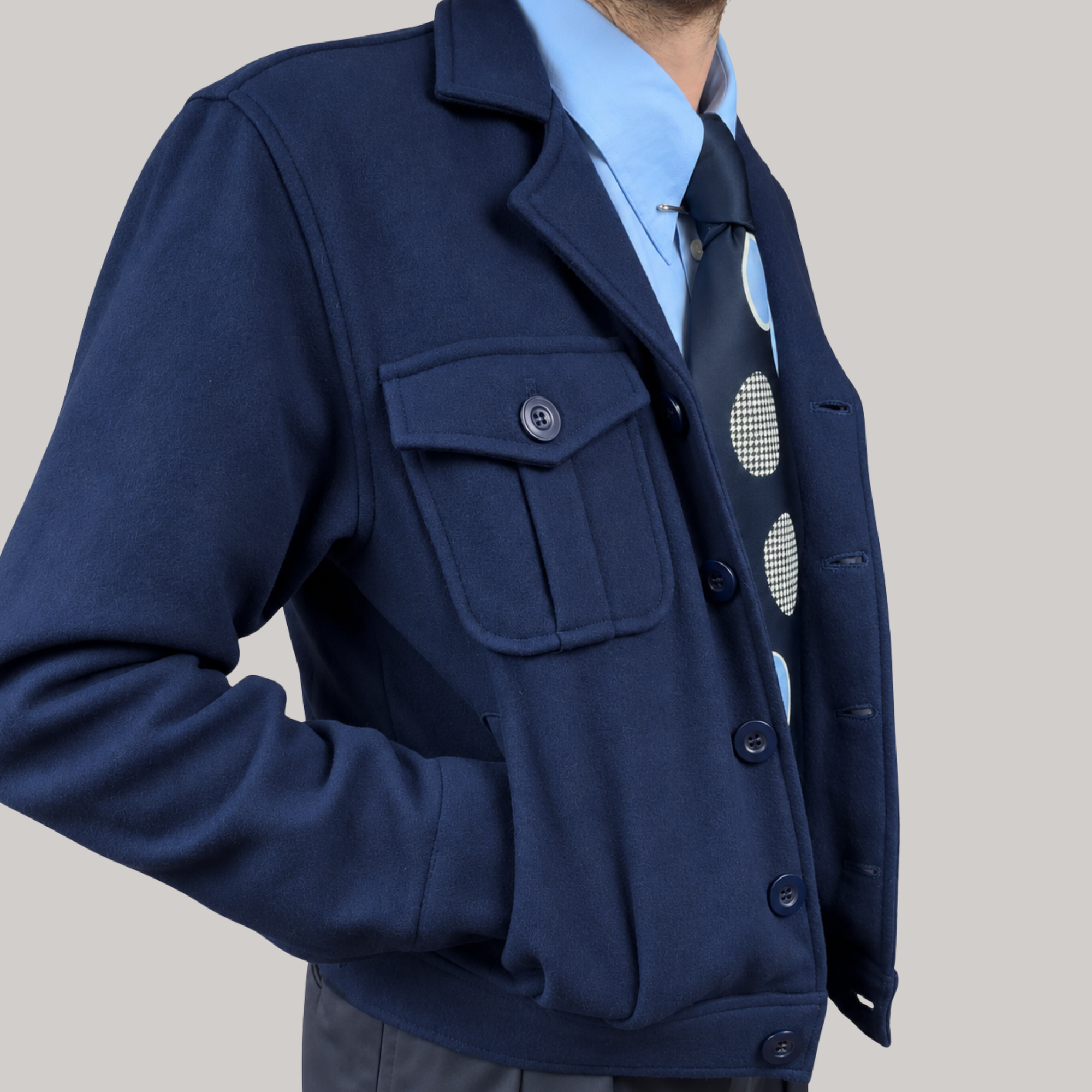 A close-up of a man wearing a navy blue wool-blend jacket over a light blue dress shirt and a dark tie with large white polka dots, set against a solid light gray background.