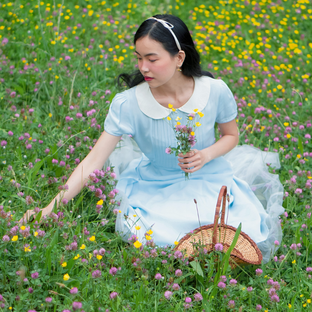 Model sits in a meadow wearing a 1940s-style shirtwaist dress in pale blue with a contrasting ceam collar. 