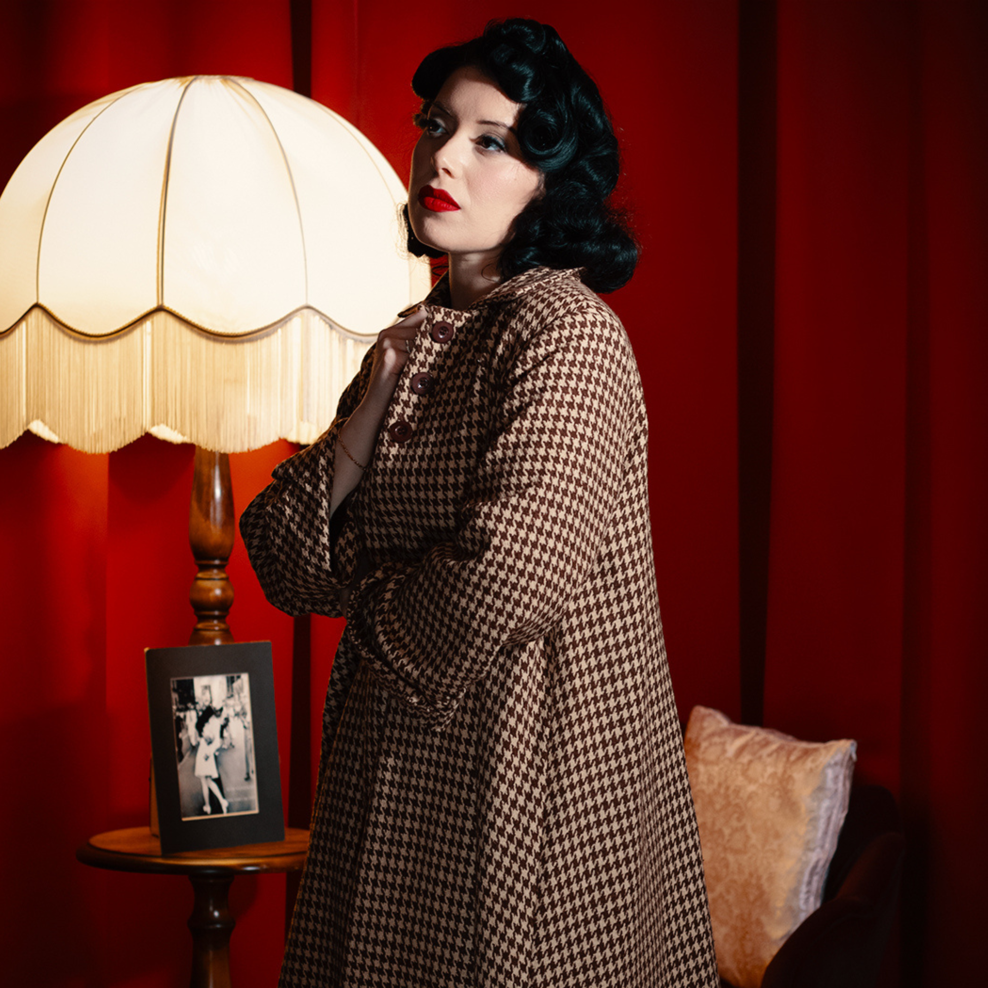 Woman in a houndstooth coat standing in a room with a lamp and framed picture on a red background