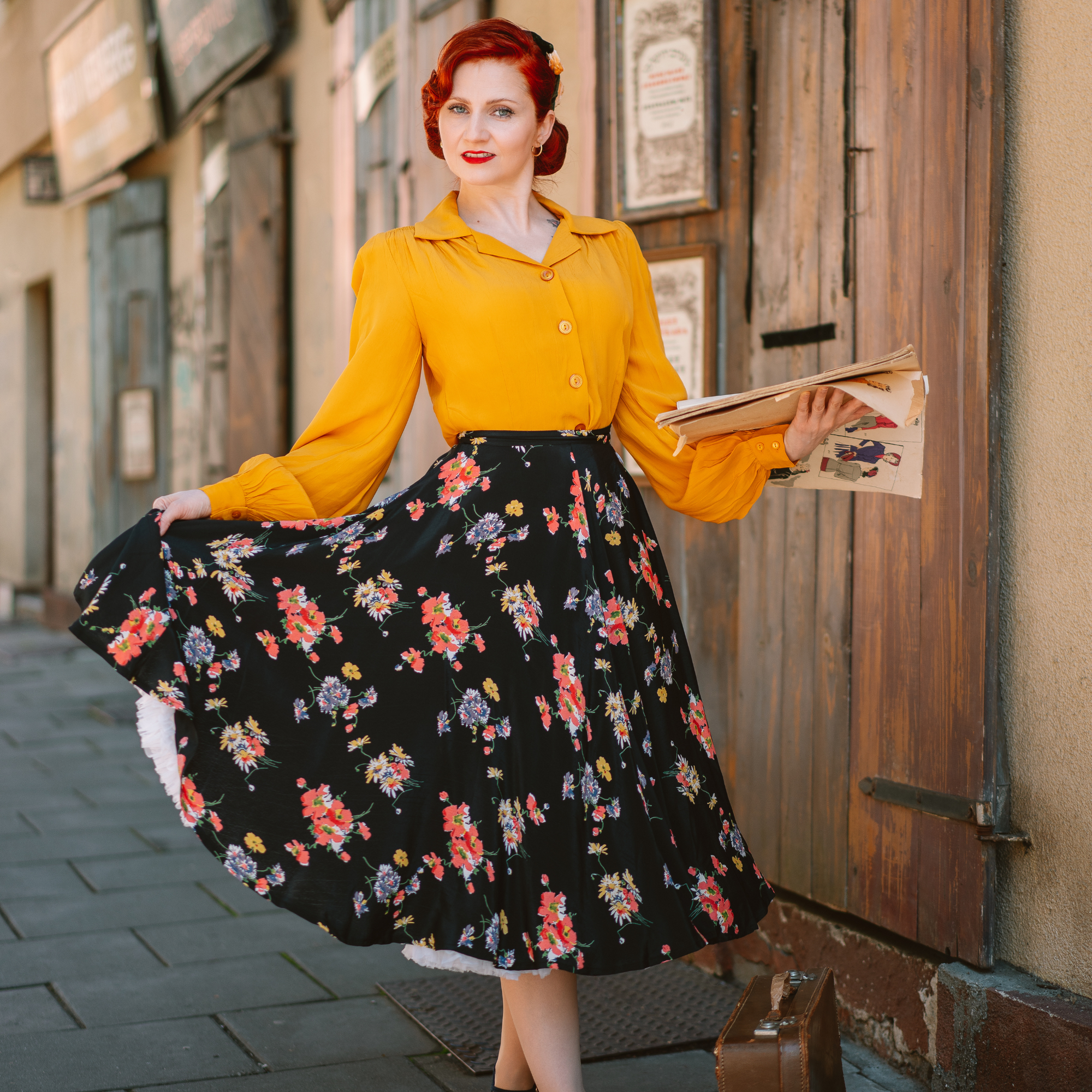 A woman wearing a mustard yellow blouse with a revere collar and long bishop sleeves, paired with a floral skirt, standing in front of a wooden door.