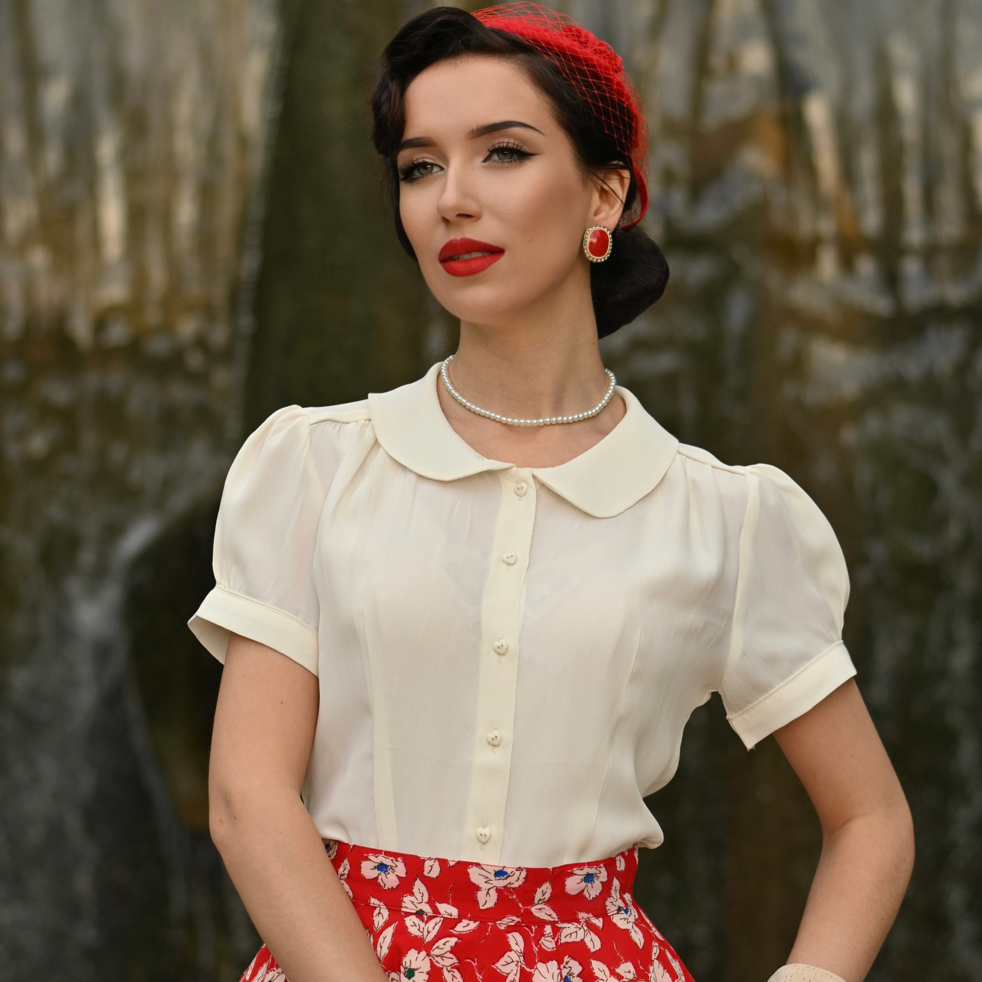 A woman wearing a cream-colored short-sleeved blouse with a Peter Pan collar and puff sleeves, paired with a red floral skirt.