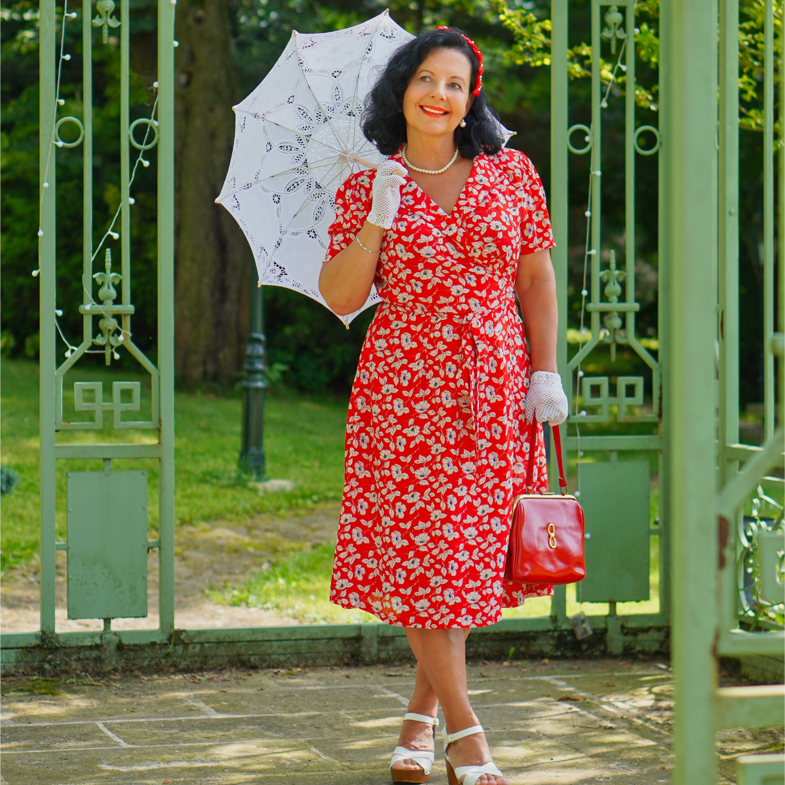 A woman standing in front of a gate, holding a white umbrella, wearing a red floral print dress with a tie at the waist, and white accessories.