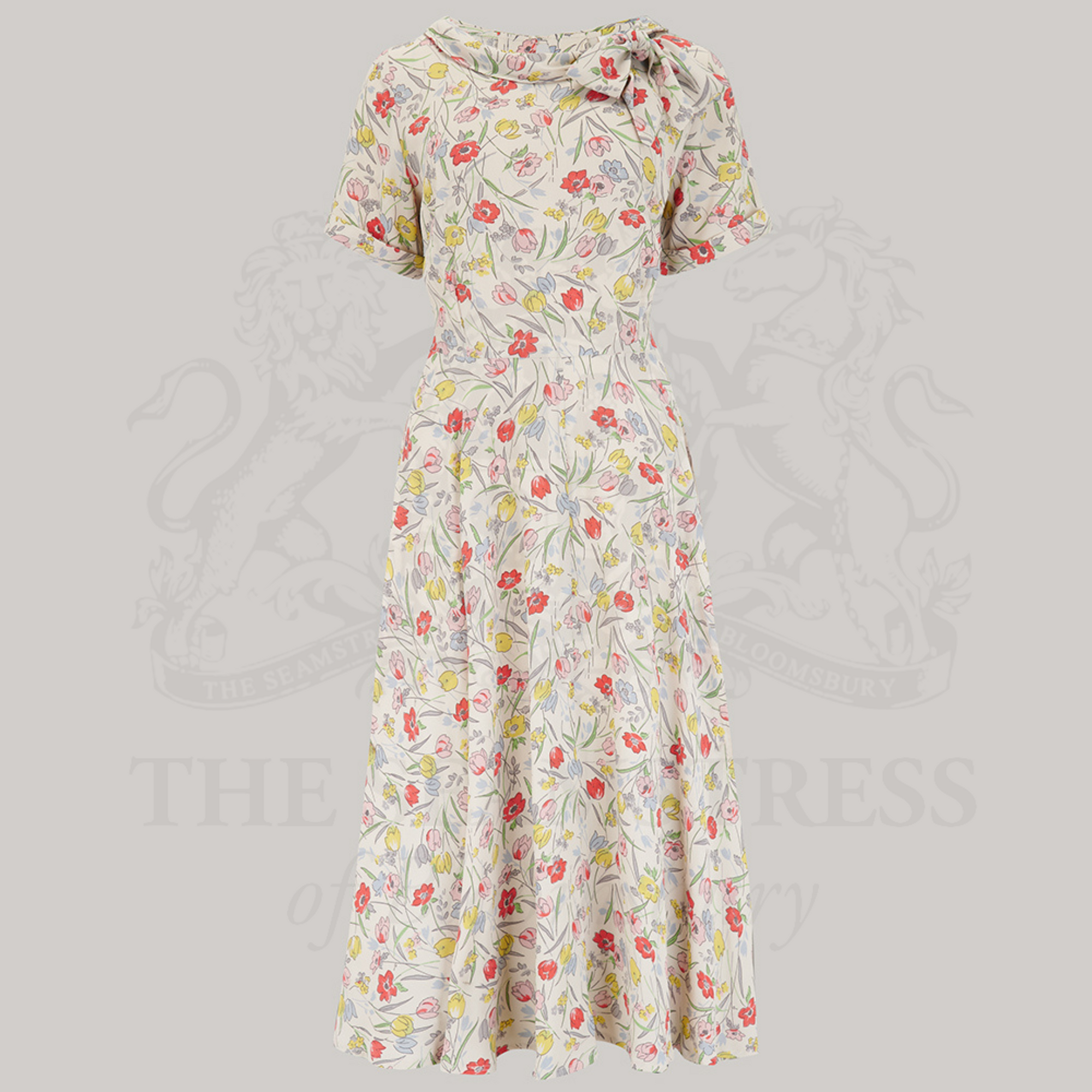 A 1940s vintage style short sleeve dress with a side tie-neck and a tie-waist belt. This dress is in a cream colour with floral patterns and has a a-line skirt and is designed to finish below the knee.