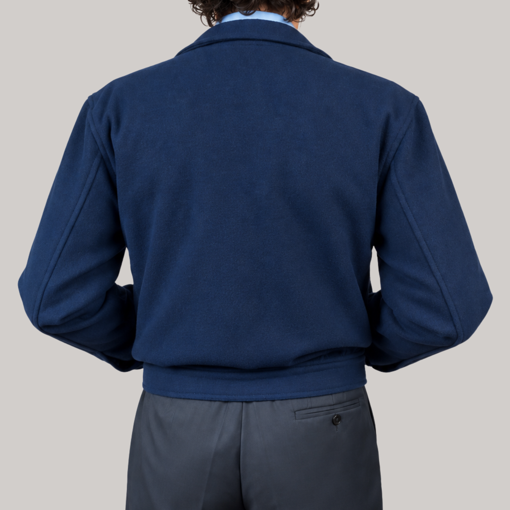 A person shown from the back wearing a navy blue wool-blend jacket over a light blue collared shirt and dark gray trousers, against a solid light gray background.