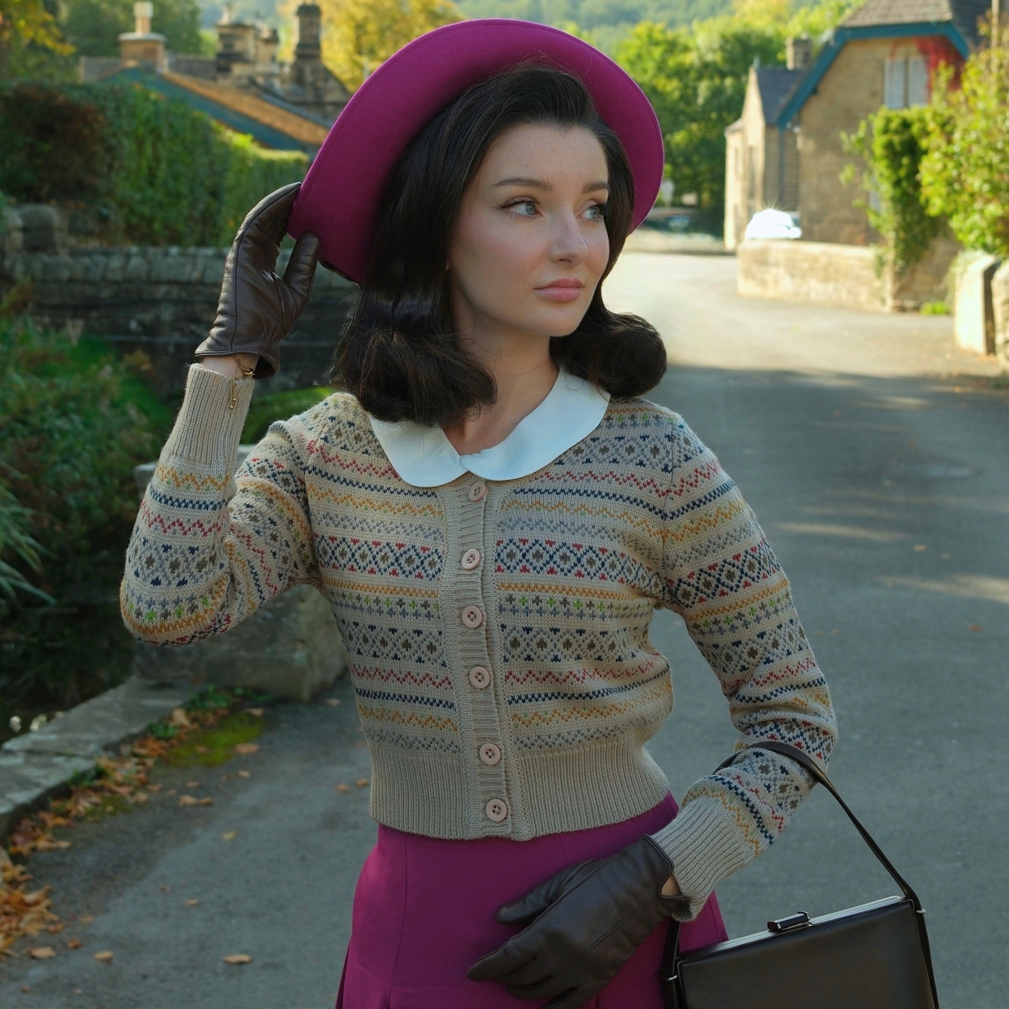 A woman stands on a country road wearing a beige Fair Isle patterned cardigan with a white Peter Pan collar, a magenta skirt, a matching magenta hat, and dark brown gloves, holding a black handbag.