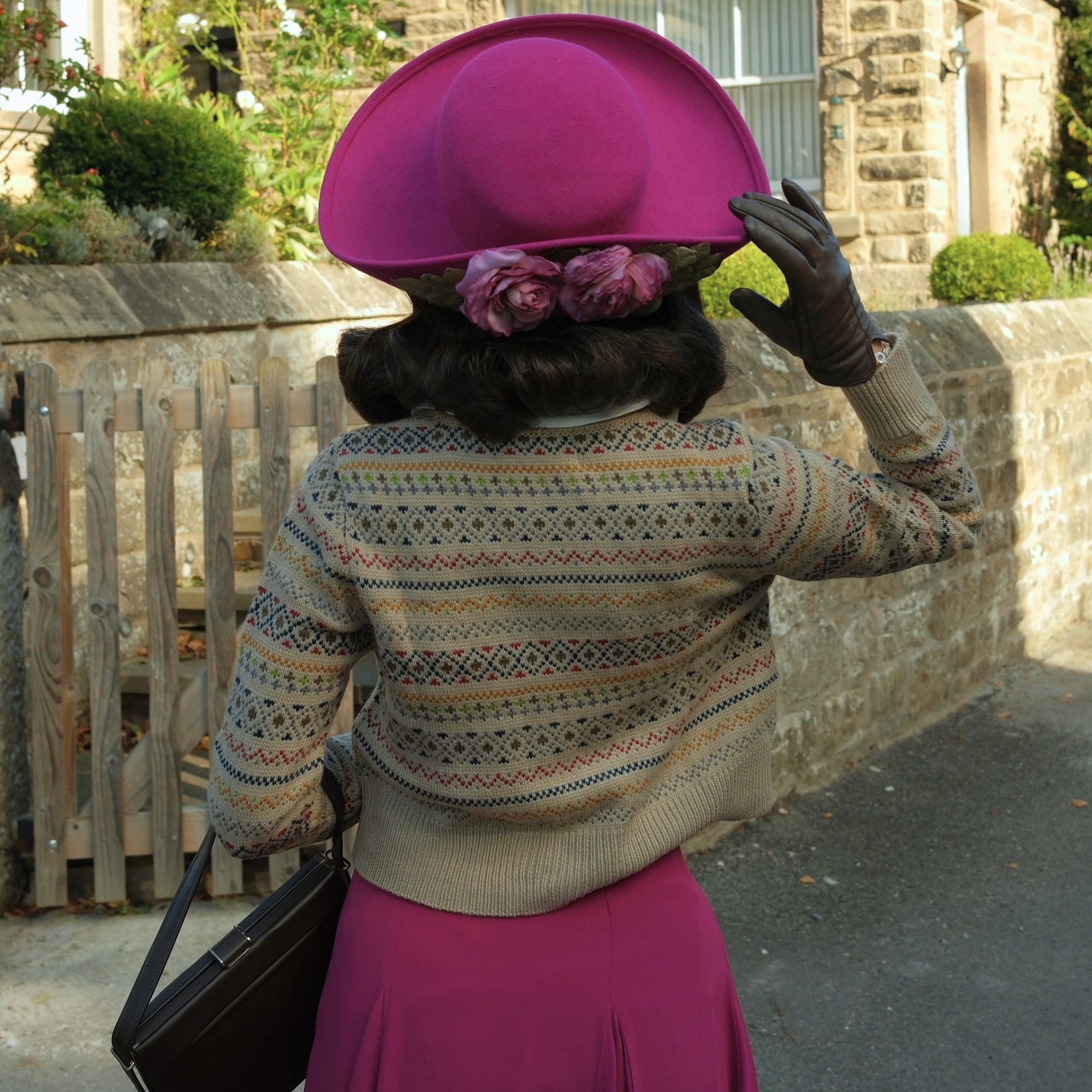 A person photographed from behind wearing a bright pink wide-brim hat with two pink flowers tucked behind the crown, a fair isle patterned knit sweater, a pink skirt, black gloves, and carrying a black handbag in an outdoor stone-path setting.