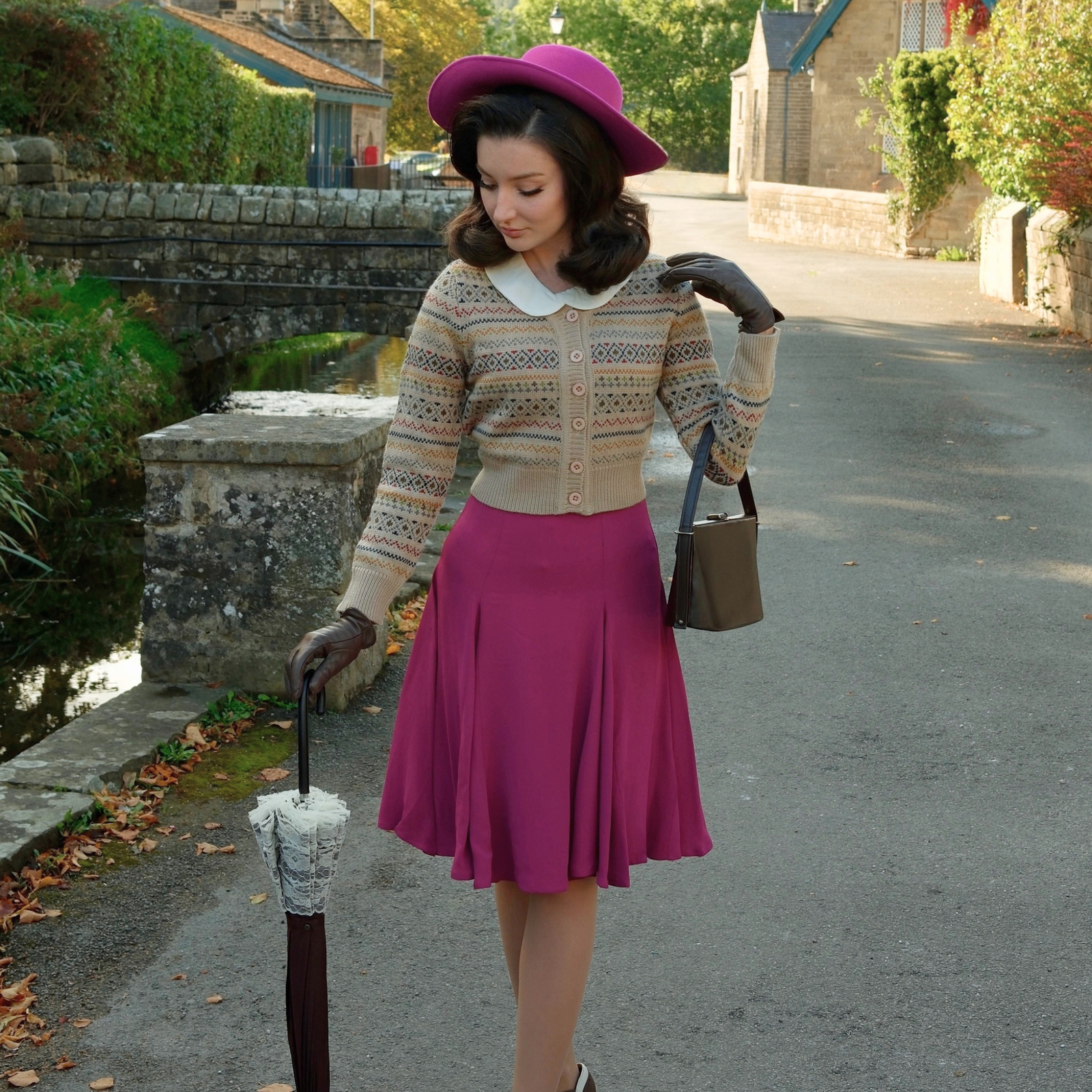 A woman stands on a quiet stone road holding a closed umbrella, wearing a bright pink wide-brim hat, a fair isle patterned knit cardigan over a cream blouse, a pink skirt, and carrying a small taupe handbag.