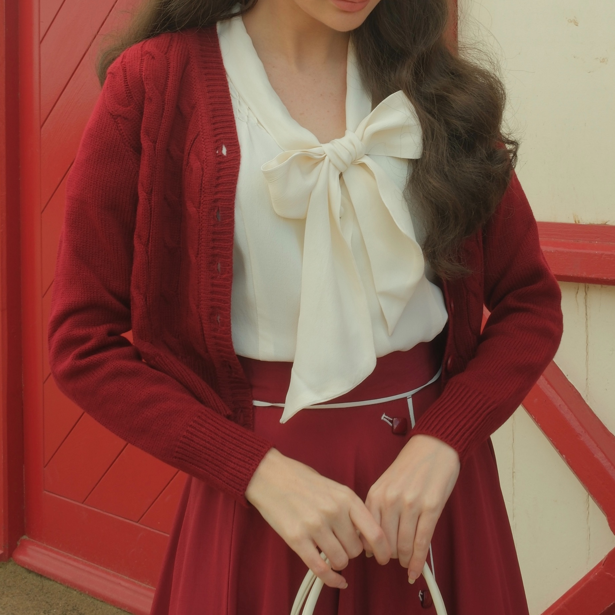 Person wearing a wine cardigan over a white blouse with a large bow, standing in front of a red door.