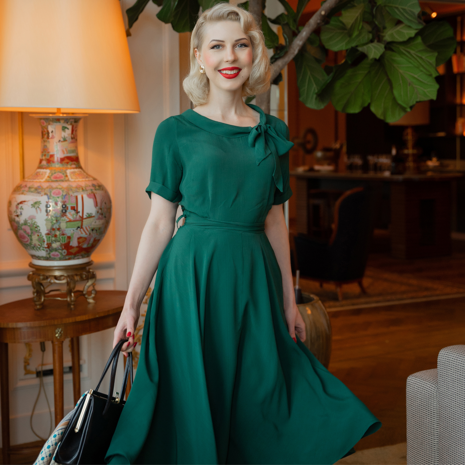 A 1940s vintage style short-sleeved dress with a side tie-neck and a tie-waist belt. This dress is dark green, has an a-line skirt, and is designed to finish below the knee.
