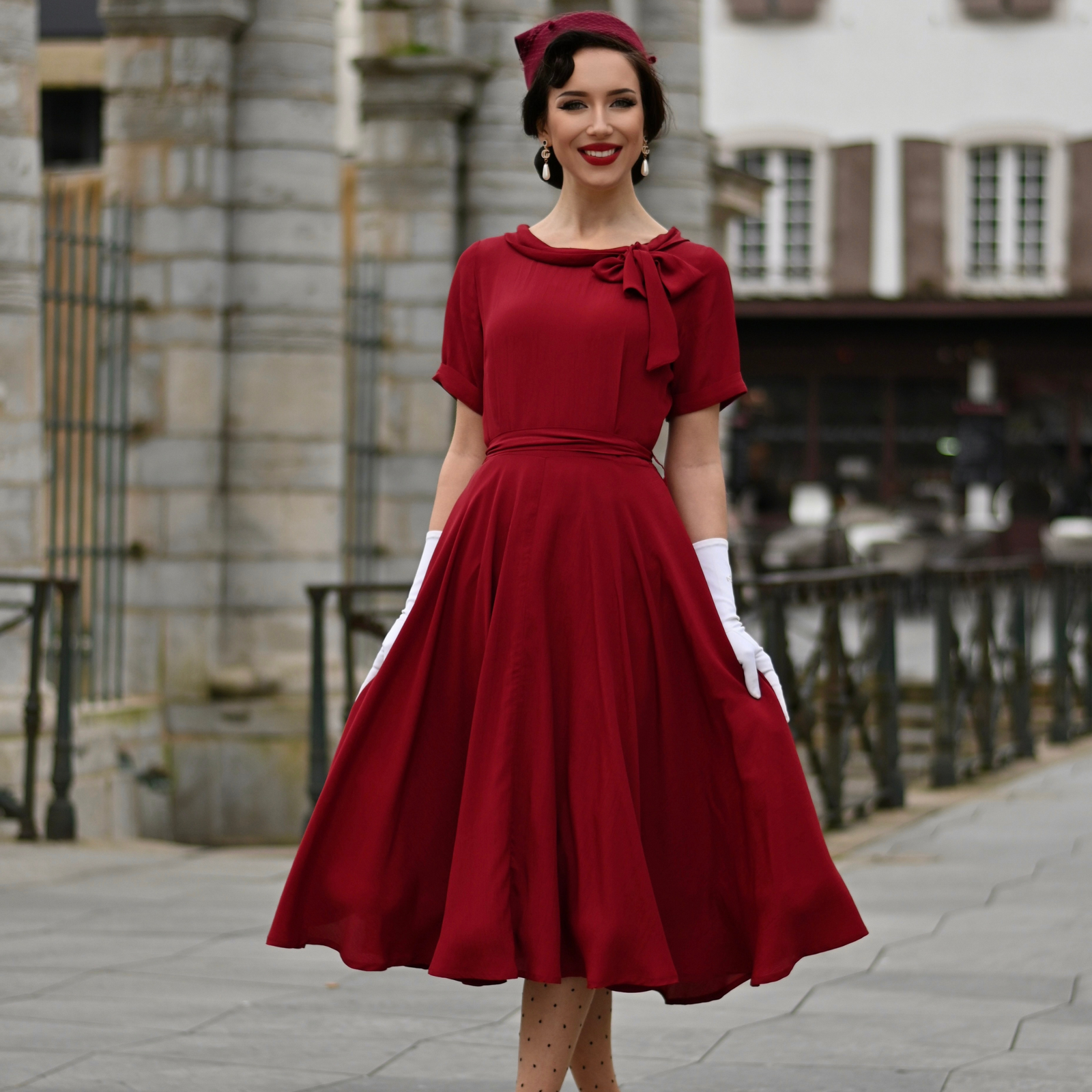Model wears a burgundy short-sleeved 1940s-style dress with a side bow collar and adjustable tie-waist belt