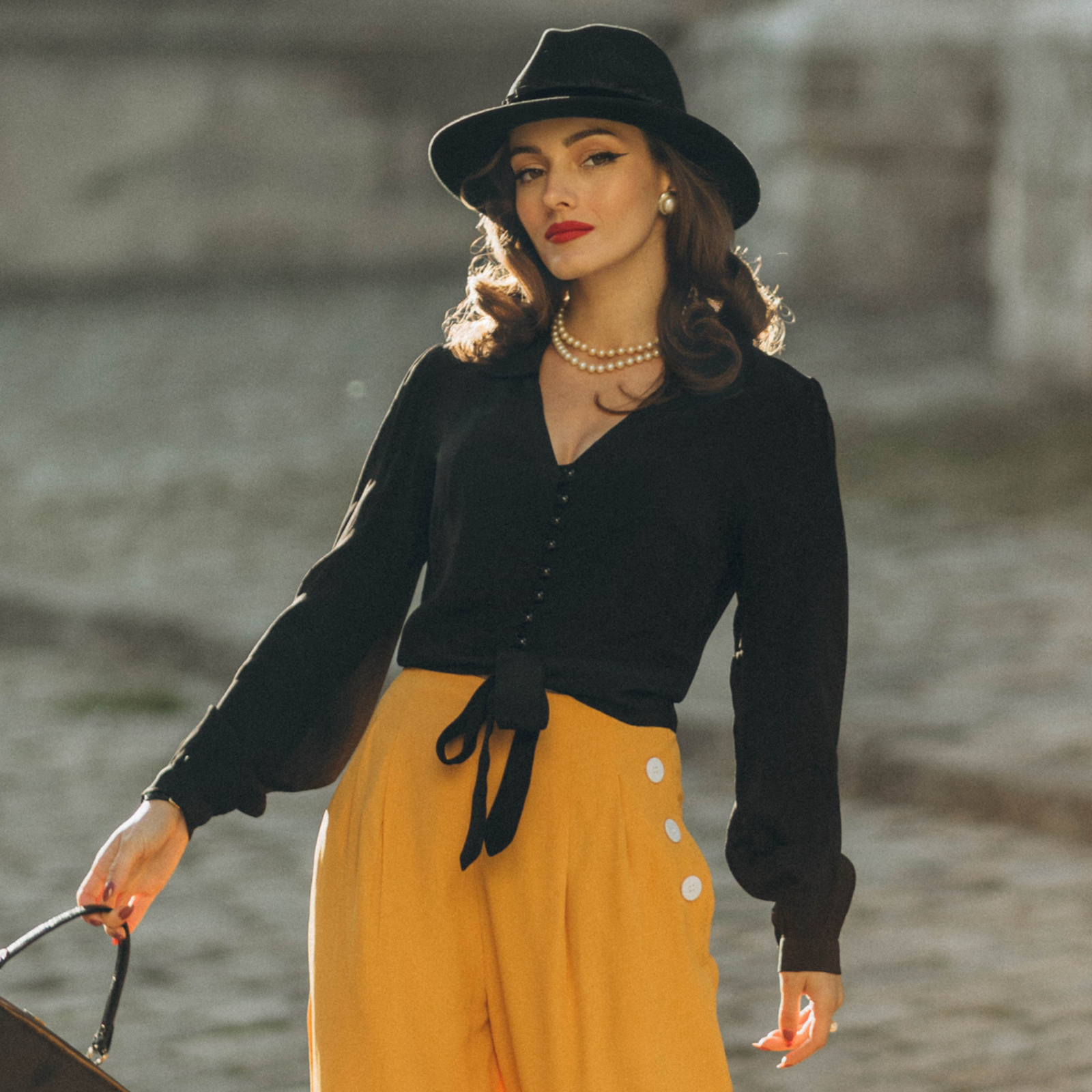 Model wears a 1940s-style black long-sleeve blouse with a v-neck, button fastenings on the front, and a waist tie. Model also wears mustard coloured wide-leg trousers
