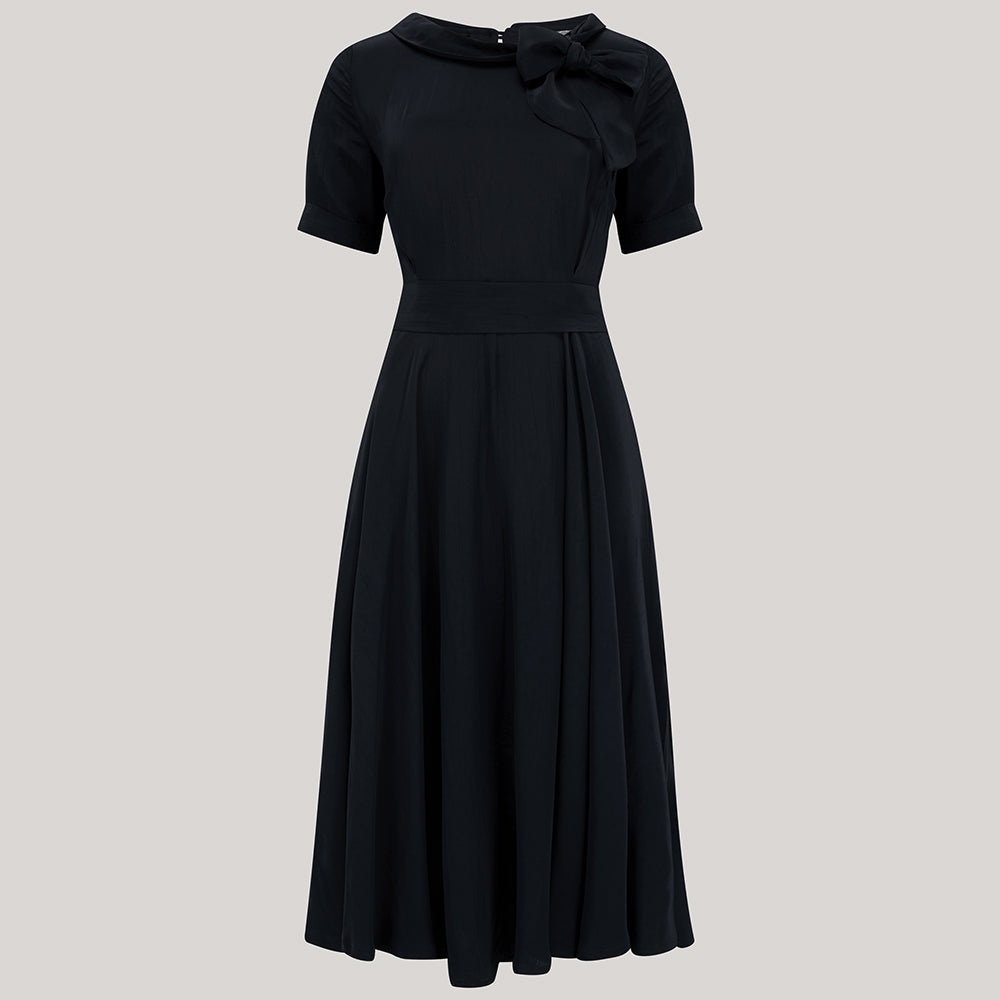 A 1940s vintage style short sleeve dress with a side tie-neck and a tie-waist belt. This dress is in a black colour and has a a-line skirt and is designed to finish below the knee.