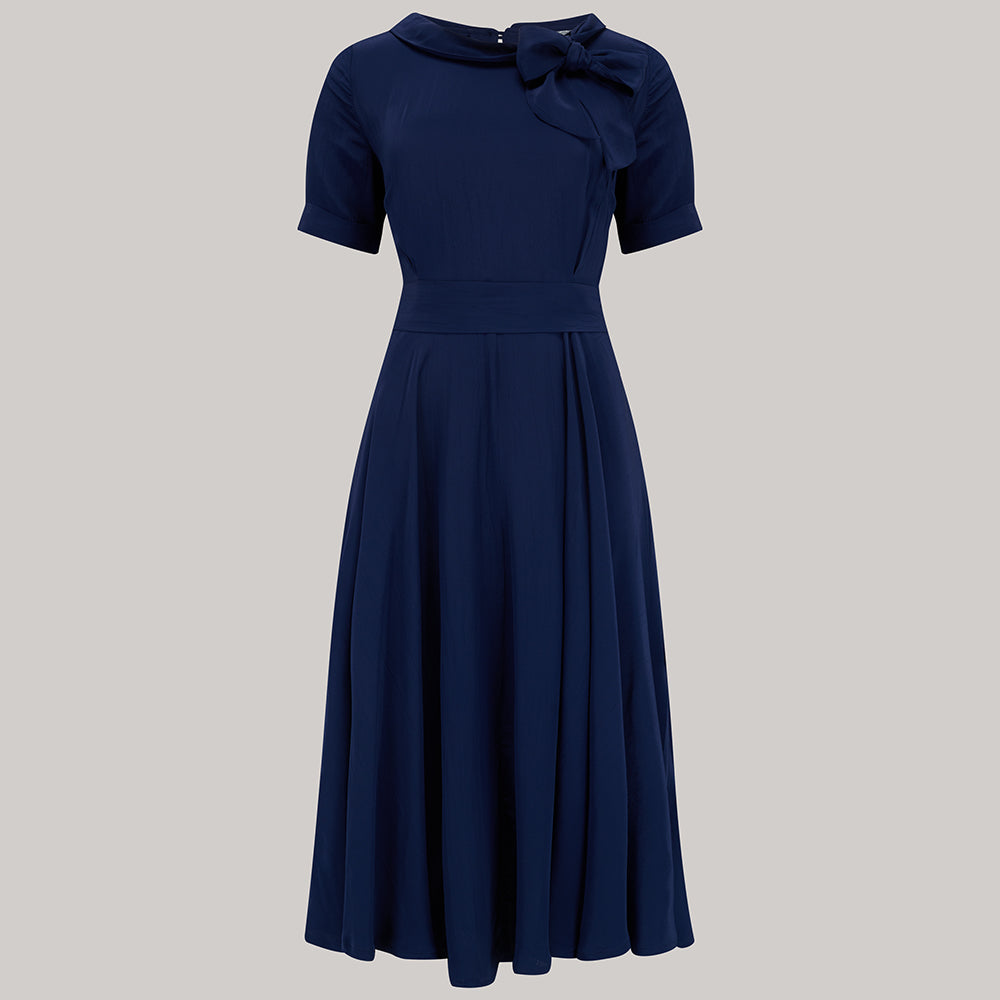 A 1940s vintage style short sleeve dress with a side tie-neck and a tie-waist belt. This dress is in a navy colour and has a a-line skirt and is designed to finish below the knee.