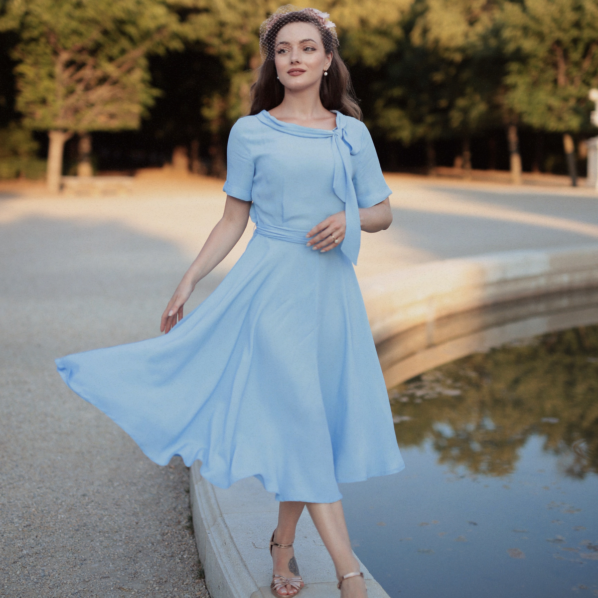 Model wears a 1940s vintage style short sleeve dress with a side tie-neck and a tie-waist belt. This dress is in a powder blue colour and has a a-line skirt and is designed to finish below the knee.