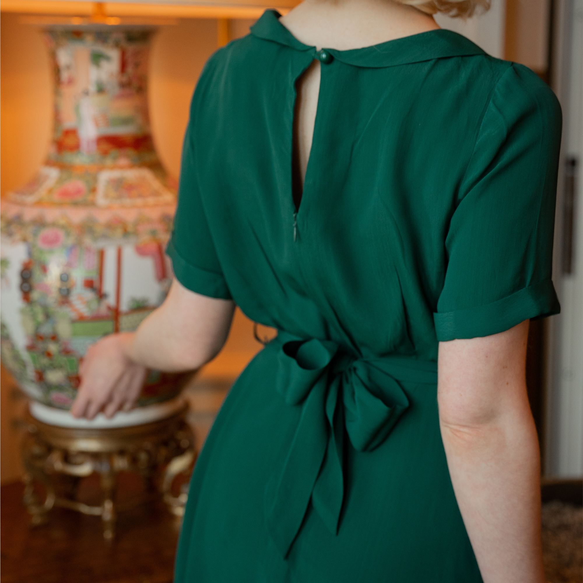 The back detailing of a vintage style green dress, showing a keyhole style on the back with a zip closure and tie-waist belt.