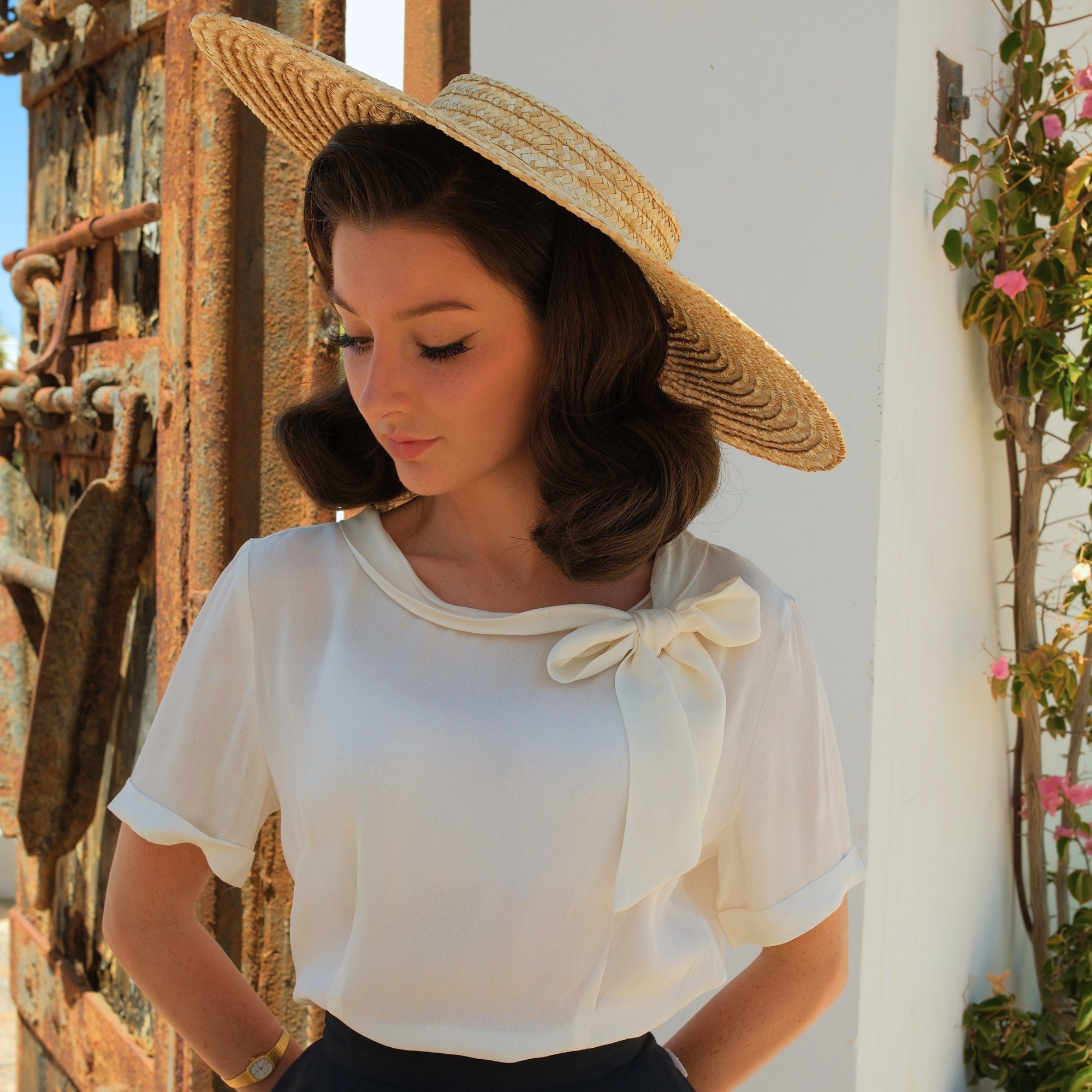 Woman wearing a white blouse with a bow and a straw hat, standing against a rustic wall.
