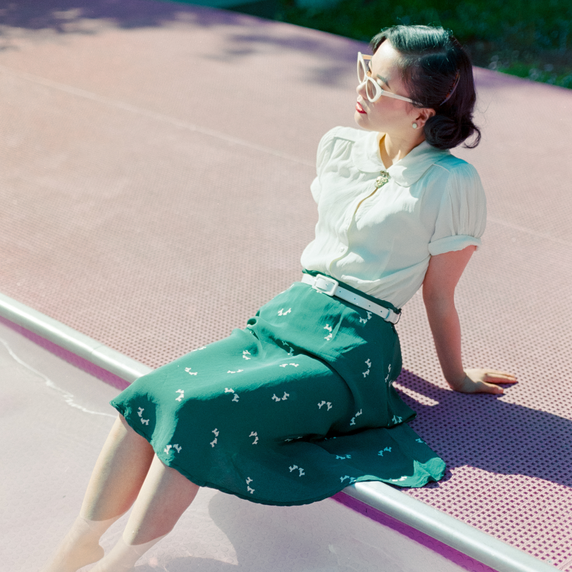 A woman wearing a white blouse and a green skirt with small white dog motifs sits on the edge of a pinkish outdoor walkway or pool deck in bright sunlight.