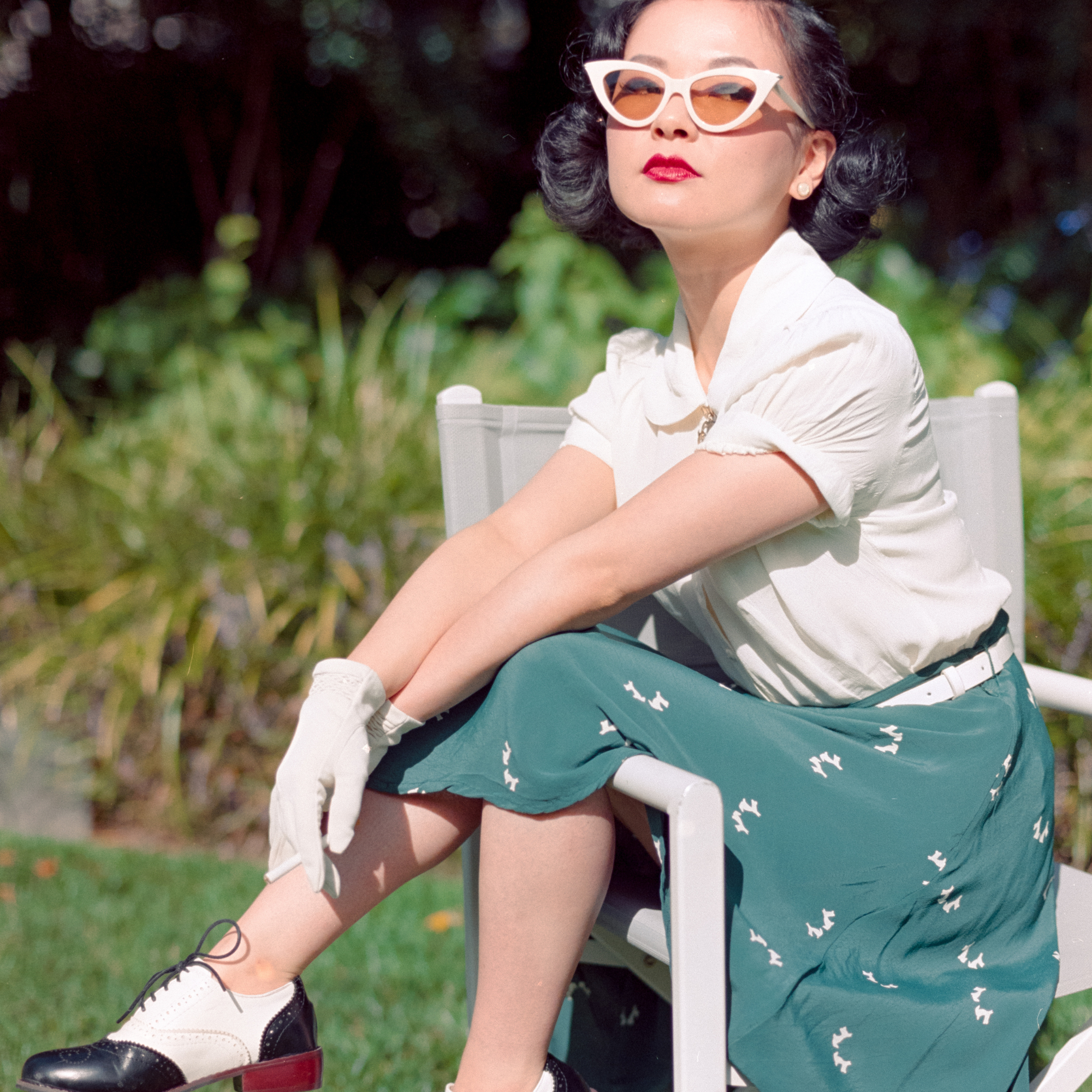 A woman in a 1940s-inspired outfit sits on a white chair outdoors. She wears a cream short-sleeved blouse, a green skirt with small white doggy prints, white gloves, and white-framed sunglasses.