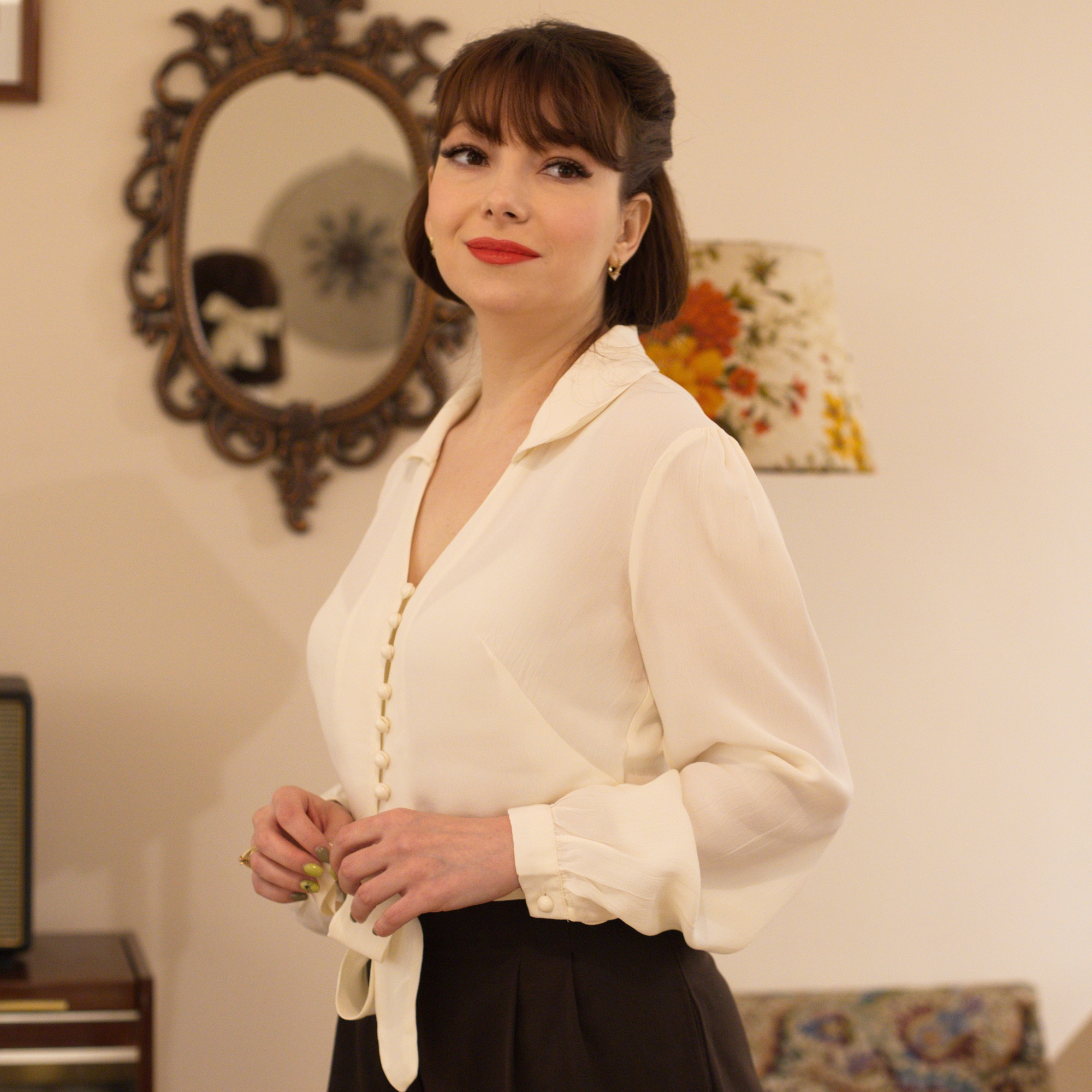 Model wears a 1940s-style cream long-sleeve blouse with a v-neck, button fastenings on the front, and a waist tie.  
