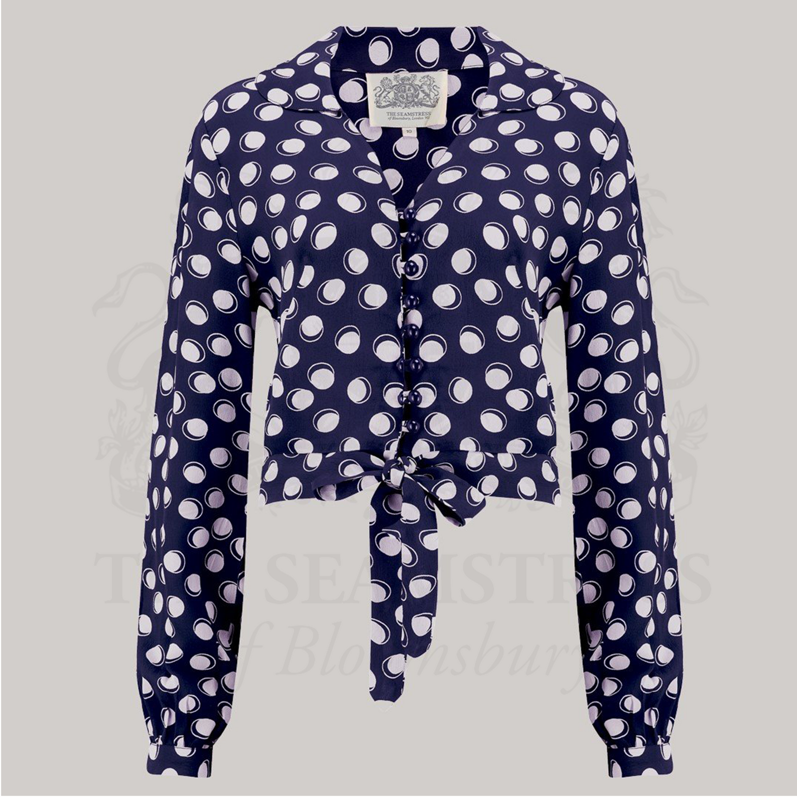 A 1940s-style long-sleeve blouse in navy with a cream polka dot pattern. The blouse has a v-neck, button fastenings on the front, and a waist tie.
