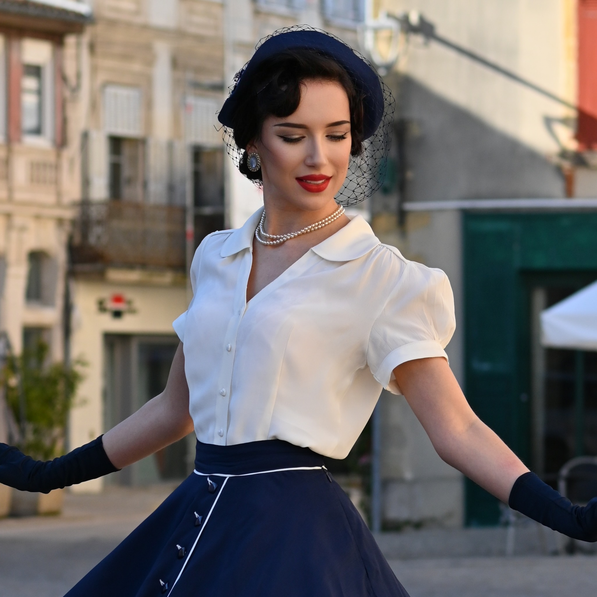 A person wearing a vintage-style cream blouse with a Peter Pan-style collar, puff sleeves, and a fitted waist, paired with a navy skirt, holding a magazine and wearing gloves.