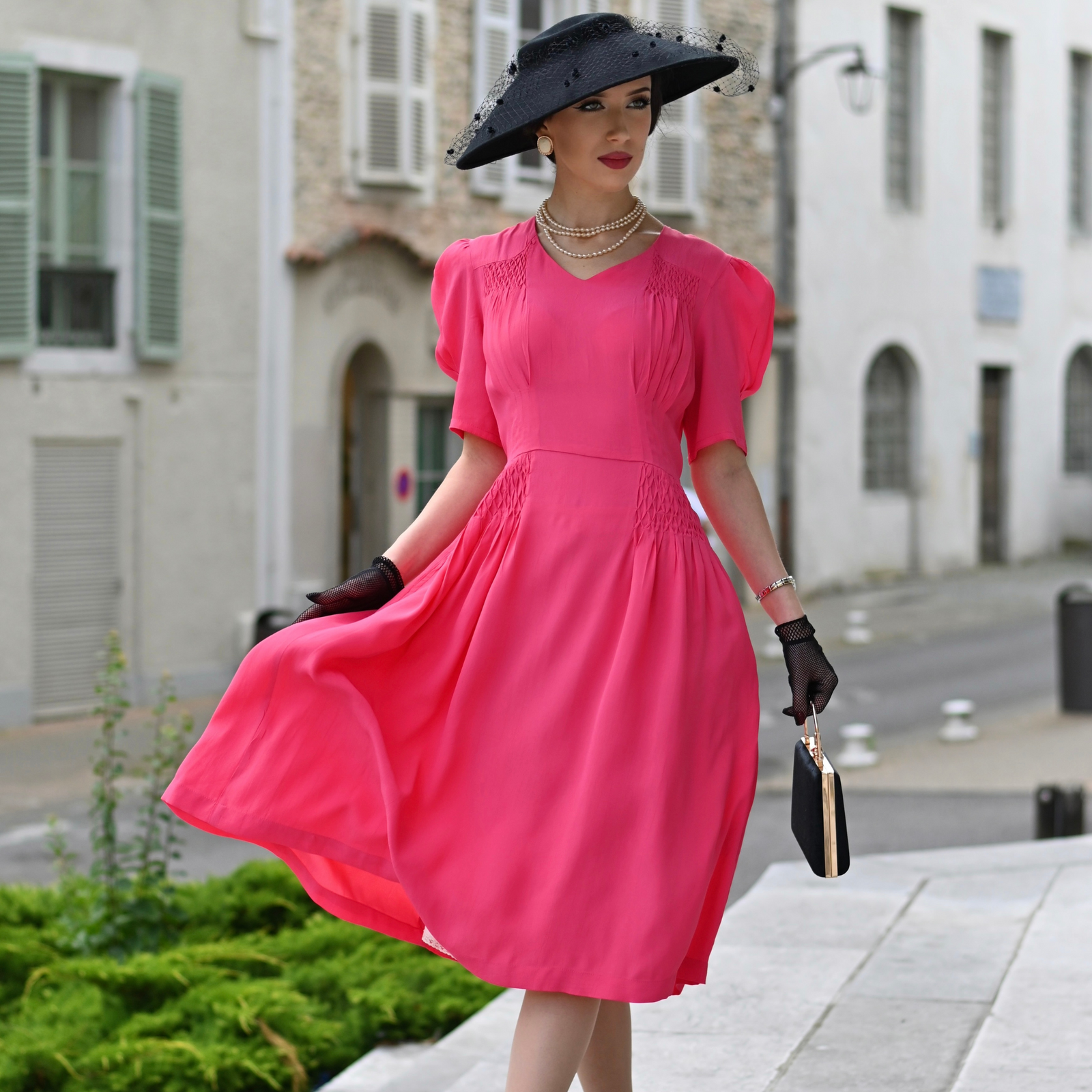 Model wears a 1940s-style raspberry pink short-sleeve blouse with a sweetheart neckline, smocking detailing on shoulders and waist, and a waist tie.  