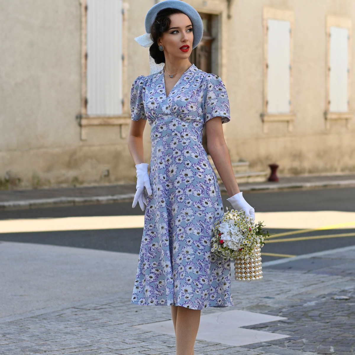 Model wears a 1940s-style tea dress in pale blue with a cream floral design, with matching cream gloves.