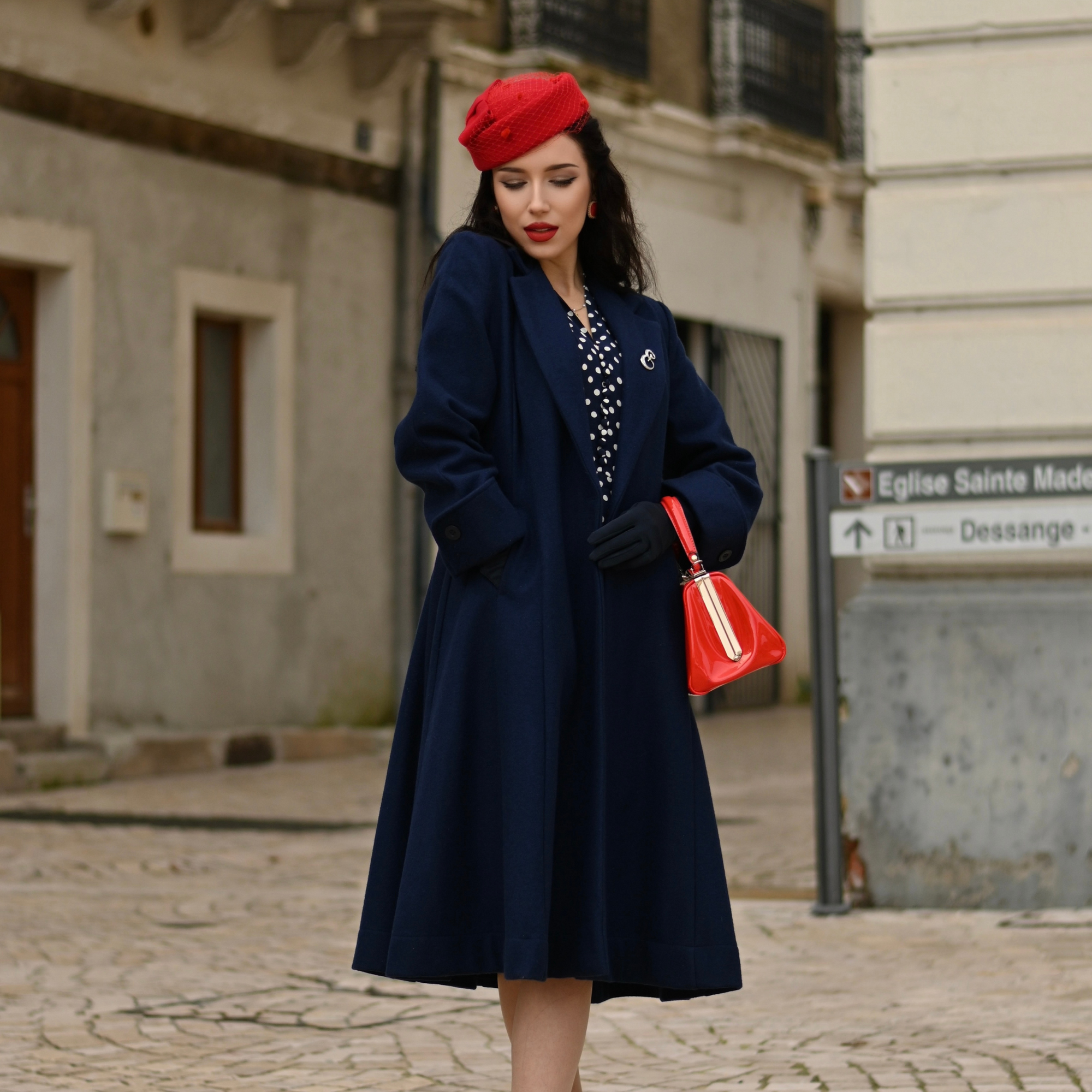 A woman wearing a navy fitted coat with side pockets, turn-back cuffs, and 4 inverted pleats in the back. She is also wearing a red beret and carrying a red handbag.