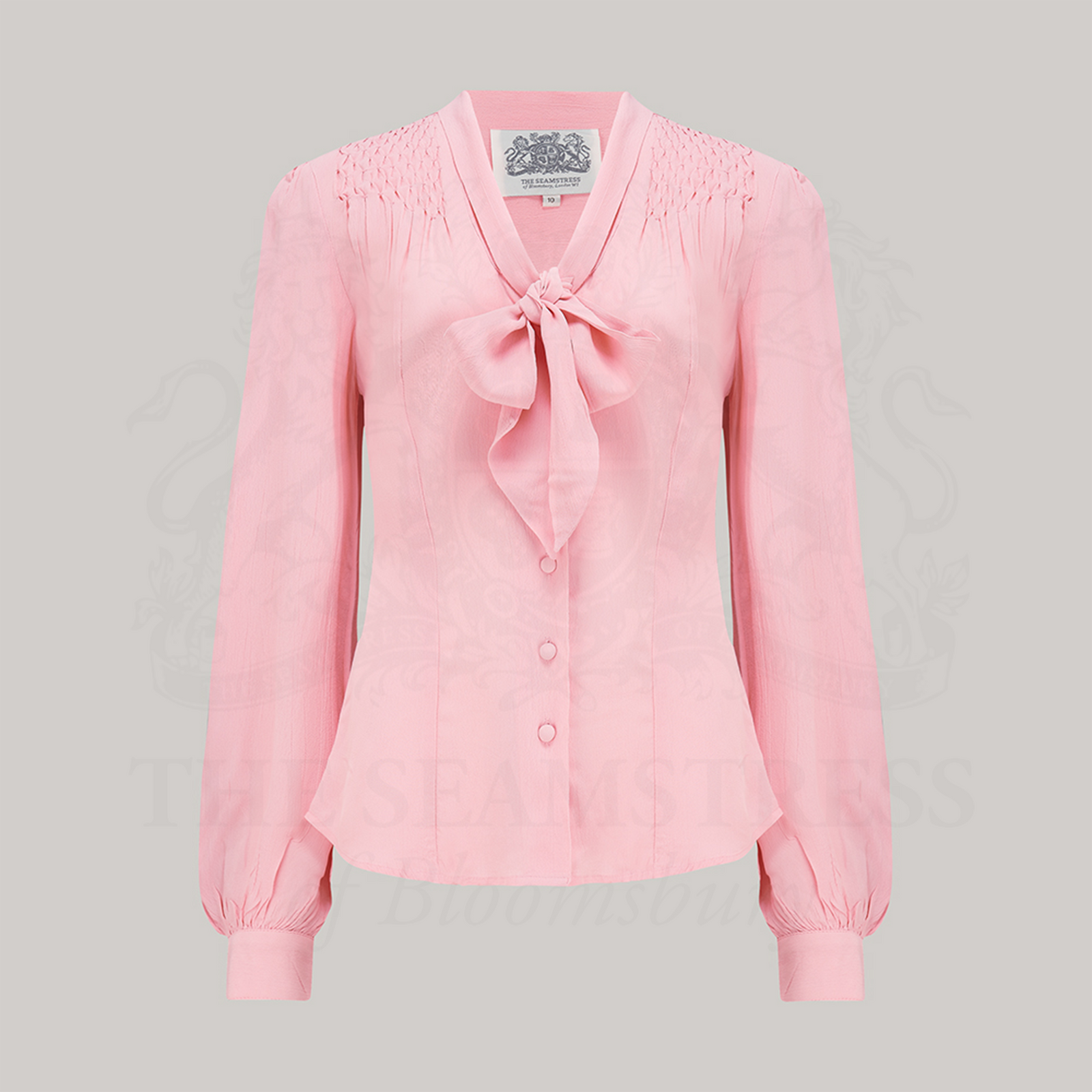 A vintage-inspired pink blouse with a pussy-bow neckline, long bishop sleeves with full cuffs, and smock stitching on shoulders displayed against a solid background.