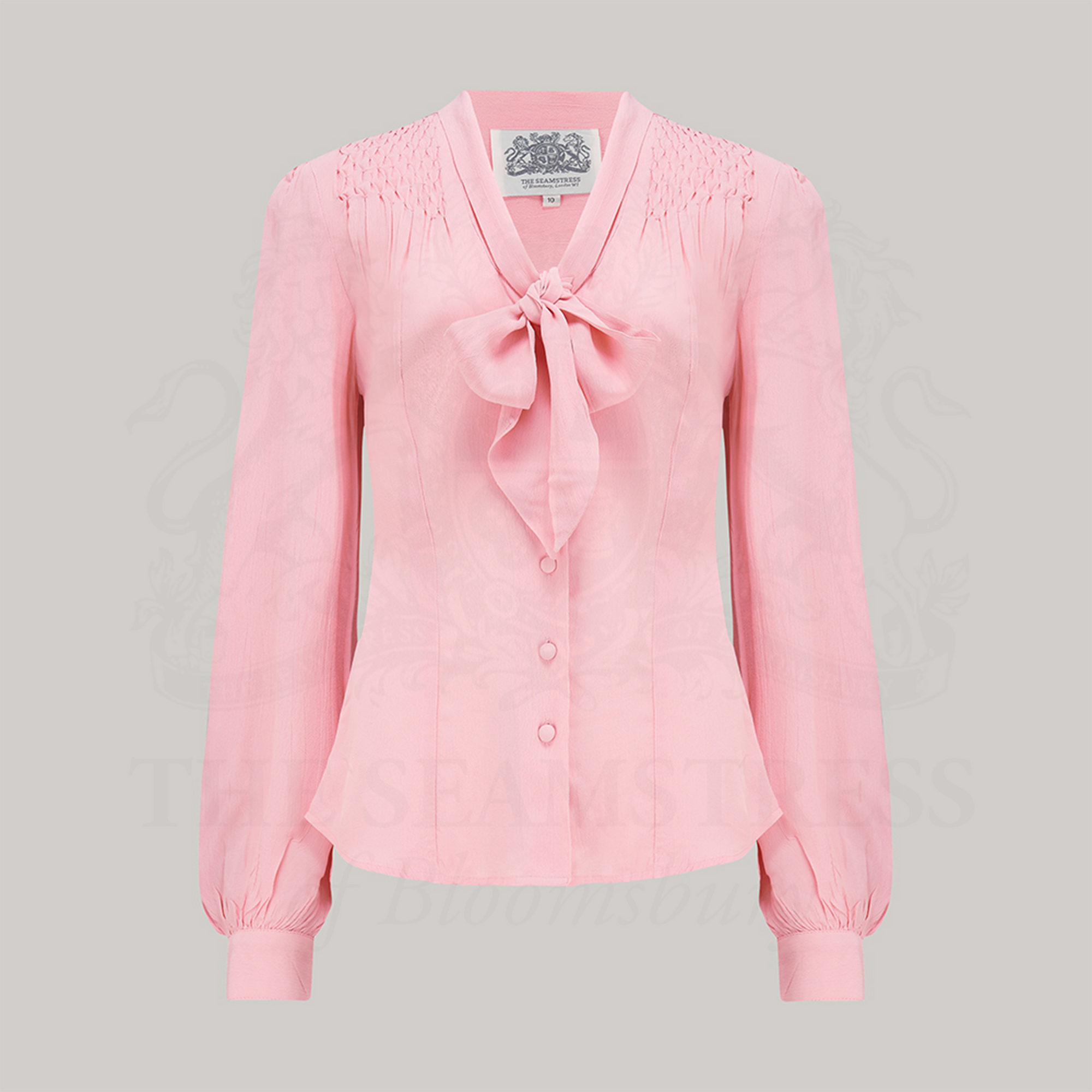 A vintage-inspired pink blouse with a pussy-bow neckline, long bishop sleeves with full cuffs, and smock stitching on shoulders displayed against a solid background.