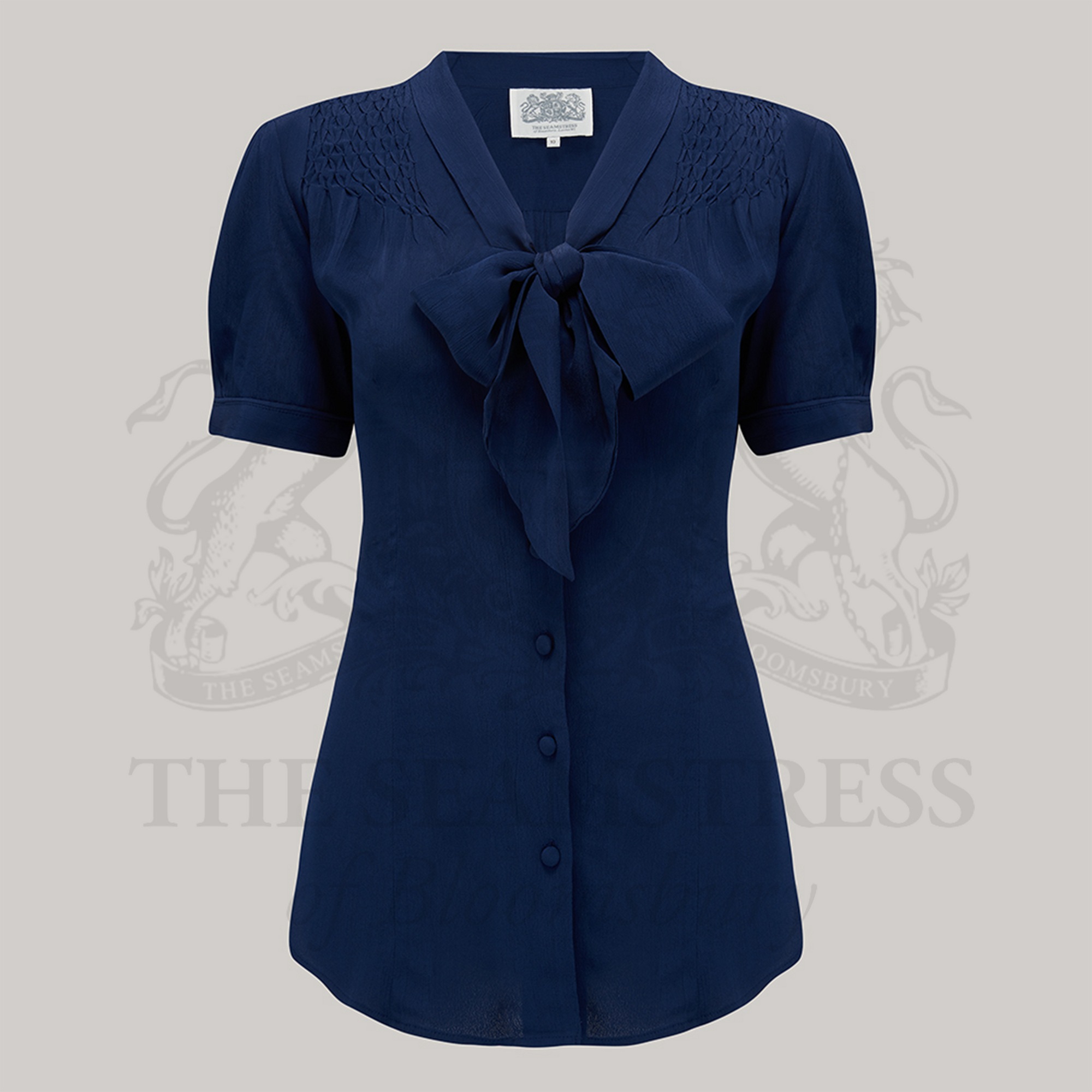 A navy-colored Eva Pussy Bow Blouse with short sleeves, smock stitching on shoulders, and button fastenings.