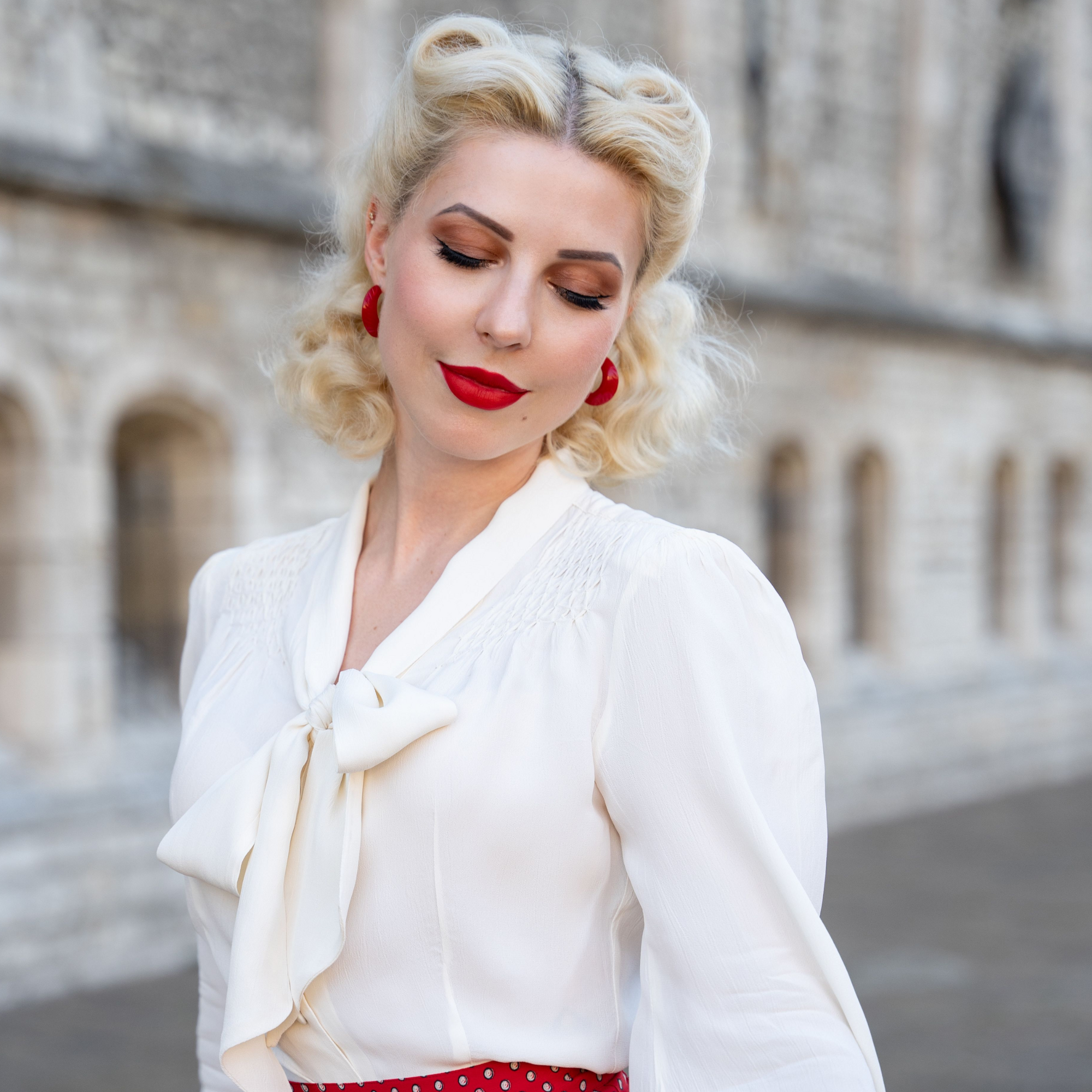 A lady wears a cream 1940s style pussybow blouse, paired with red lipstick and matching accessories