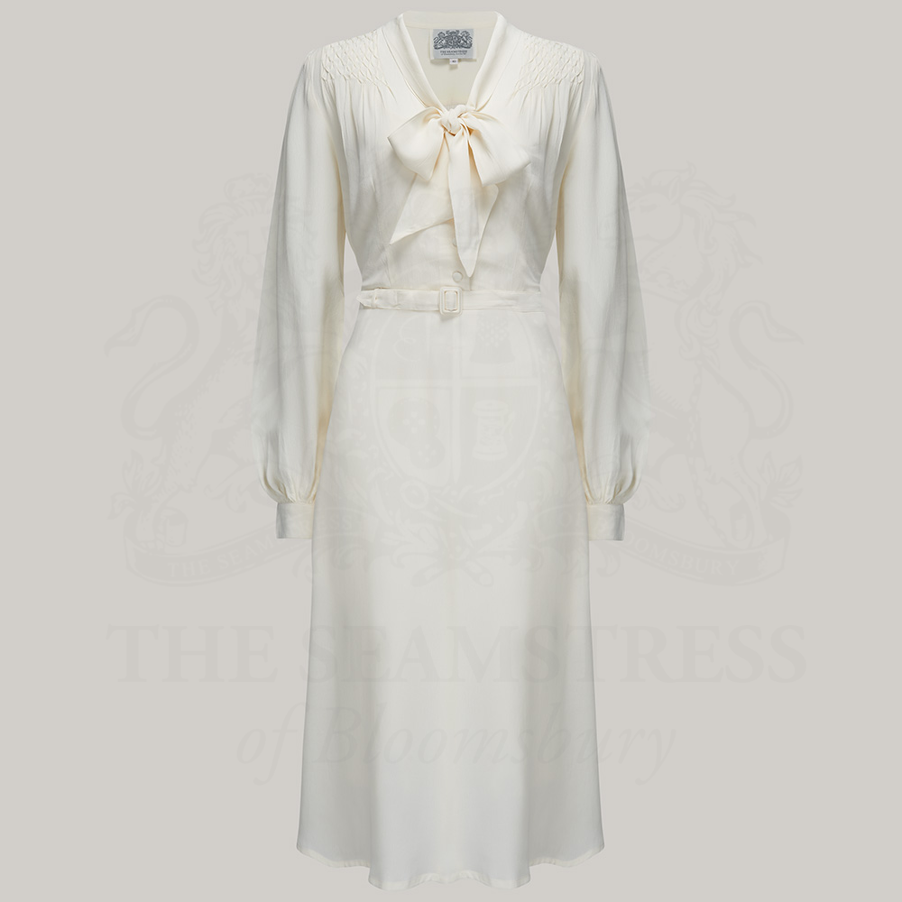 A vintage-inspired cream dress with a pussy bow neckline, long sleeves, and smock stitching on the shoulders. The dress features button fastenings, a side zip, waist belt, and side-seam pockets, paired with a complimentary slip.