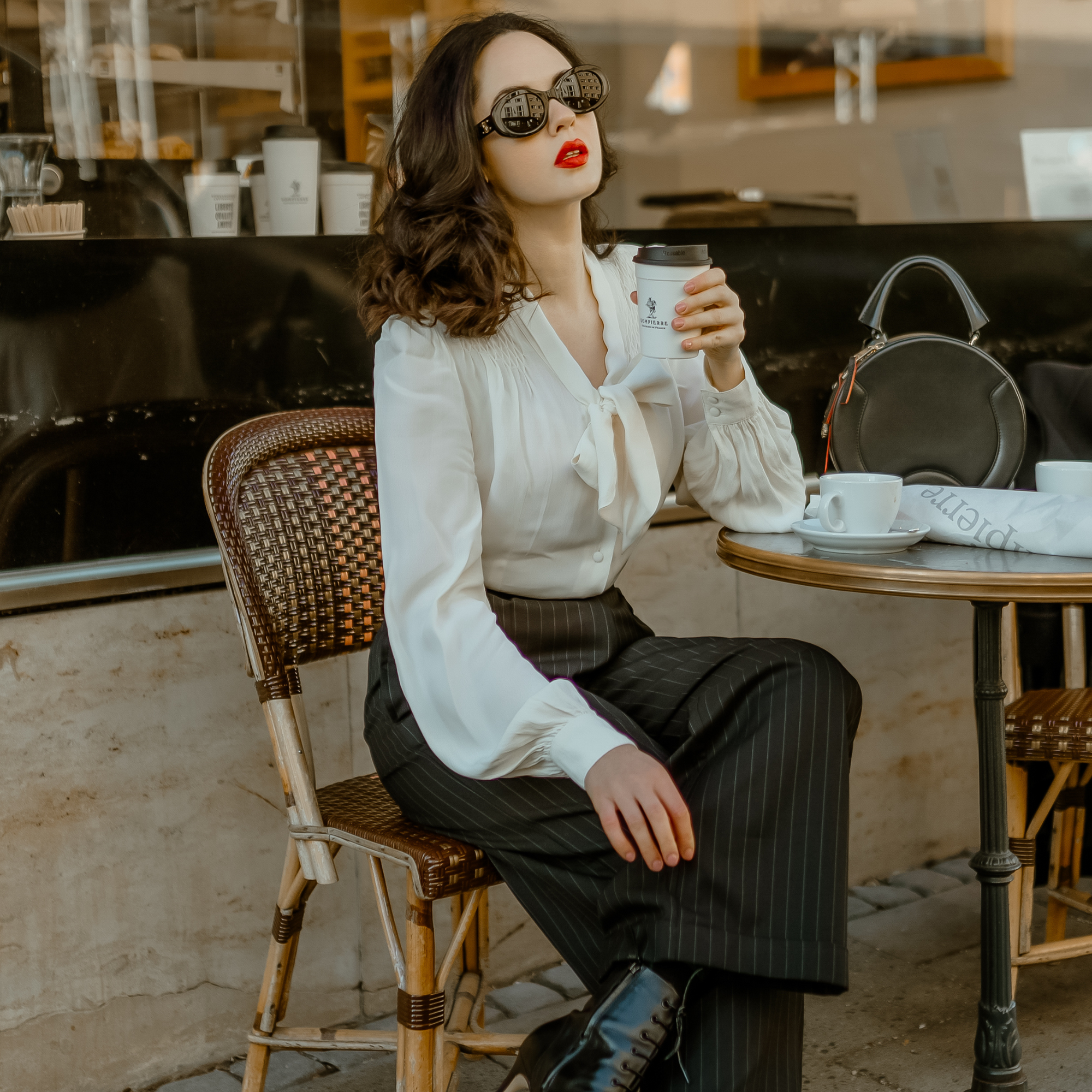 A model wearing a 40s style cream pussy-bow blouse with long bishop sleeves. She wears tailored black pinstripe trousers, paired with black sunglasses