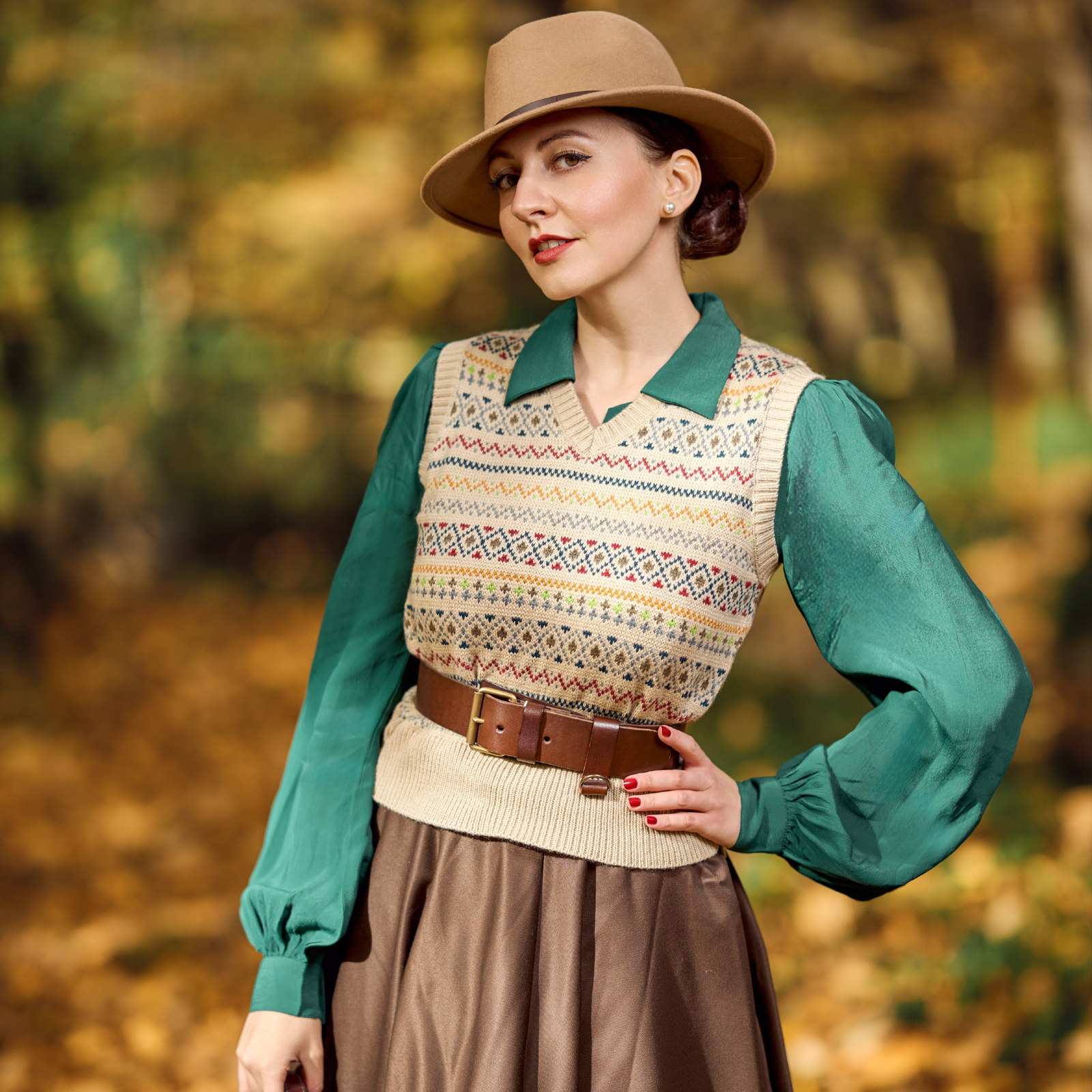 Woman in a green blouse and patterned vest with a brown hat and skirt standing in an autumn setting.