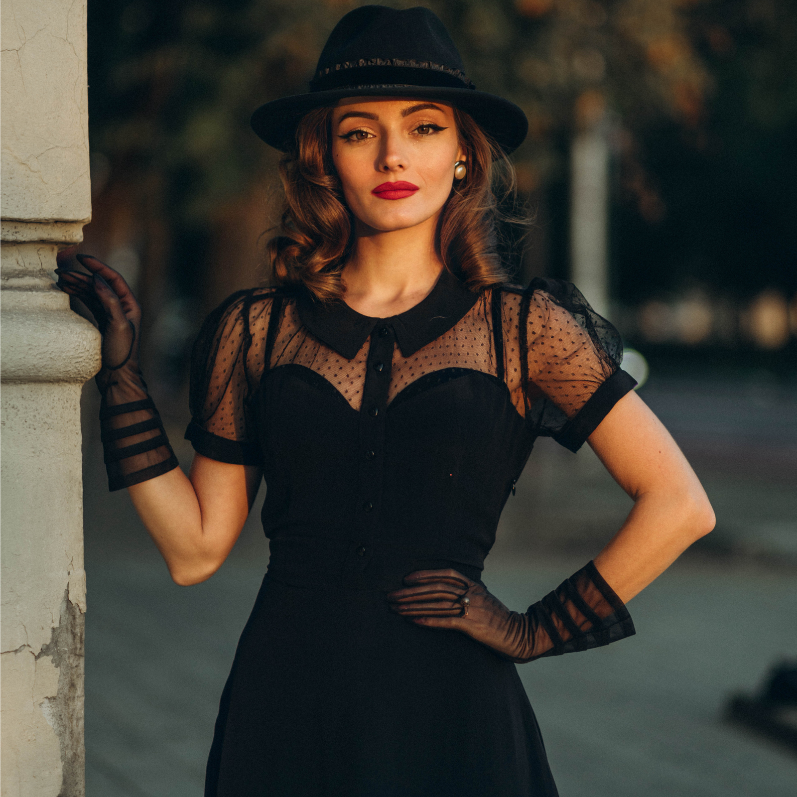 A woman wearing a black dress with a 1940s-inspired pointed flat collar, short sleeves, and net detailing. She is also wearing a tie-waist belt and heart-shaped buttons, accessorized with a hat and gloves.