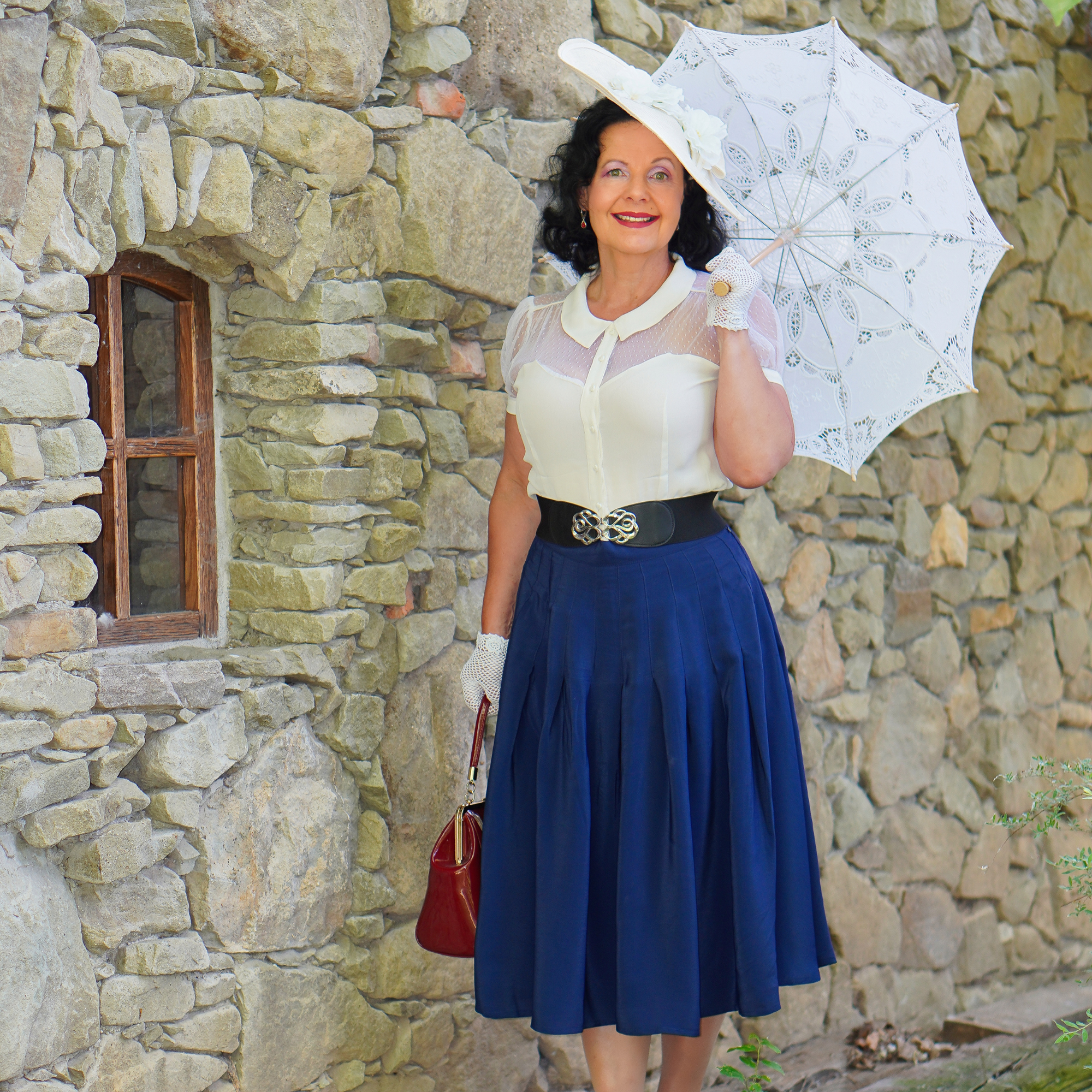 Woman in vintage attire holding a white umbrella against a stone wall.