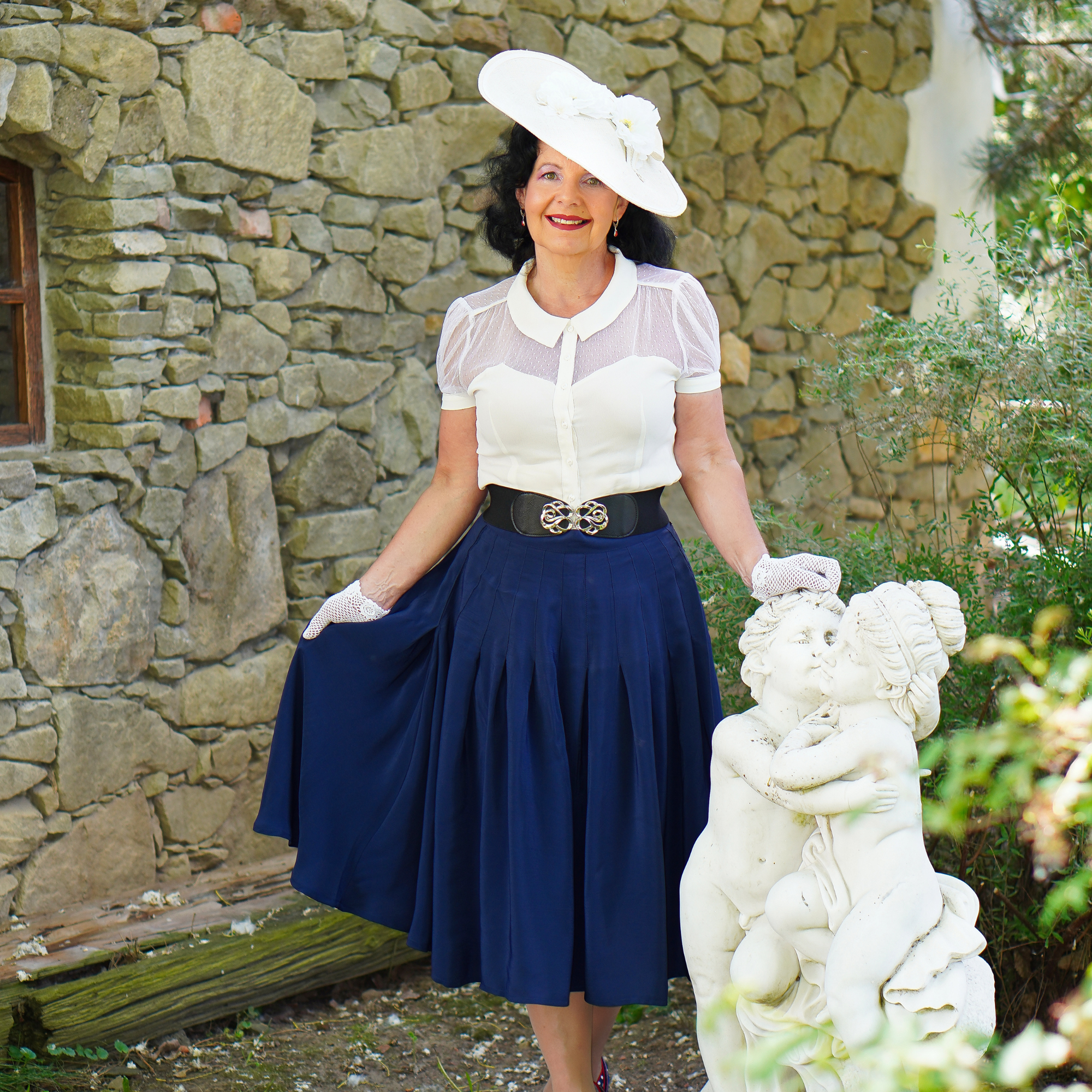 Woman in a white blouse and navy skirt standing next to a stone wall and statue.